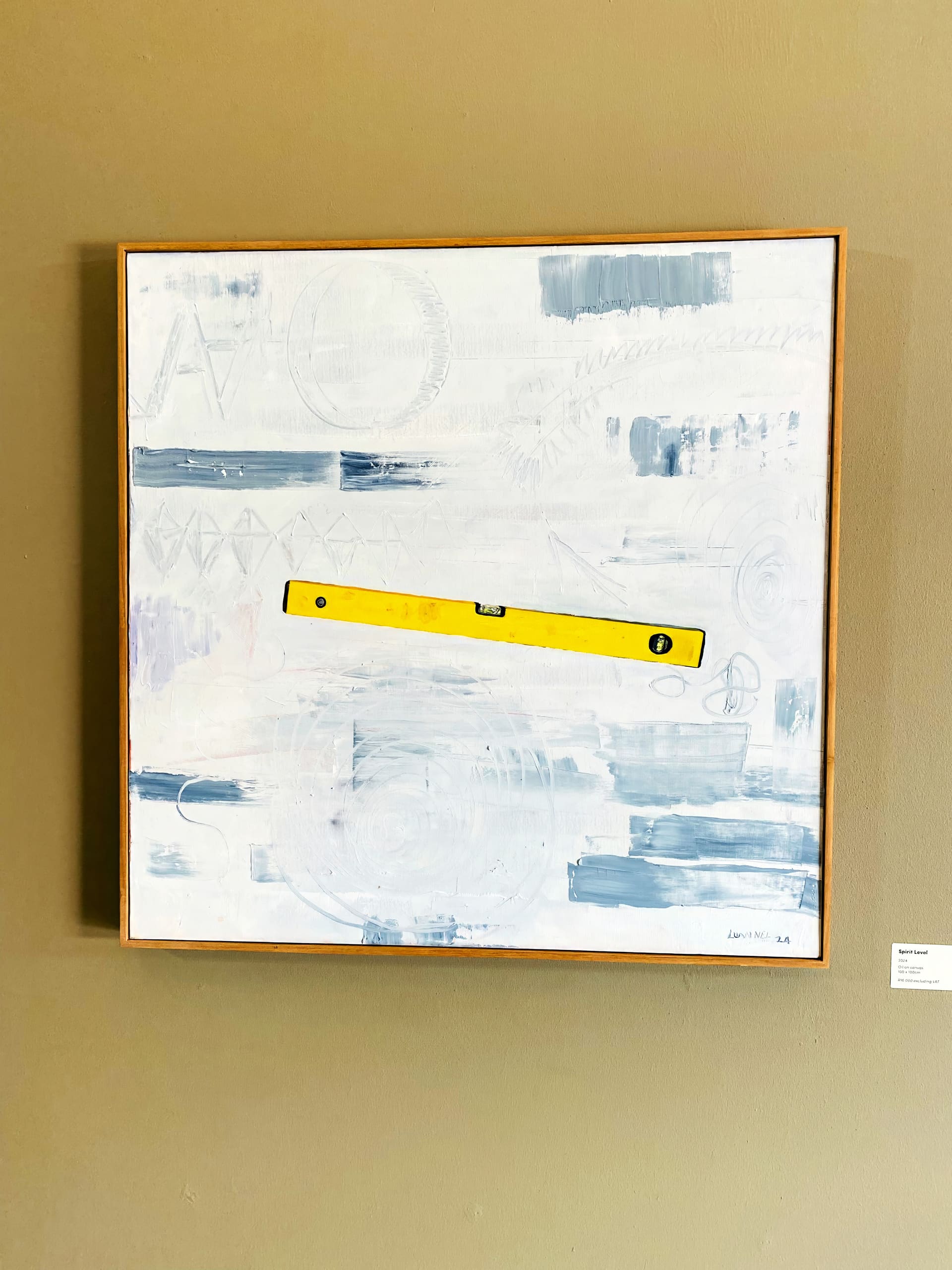 A framed abstract painting with a pale, textured background in white and light bluish tones. A bright yellow spirit level is horizontally centred across the canvas. The piece hangs on a beige wall, with a small, white placard visible at the bottom right.