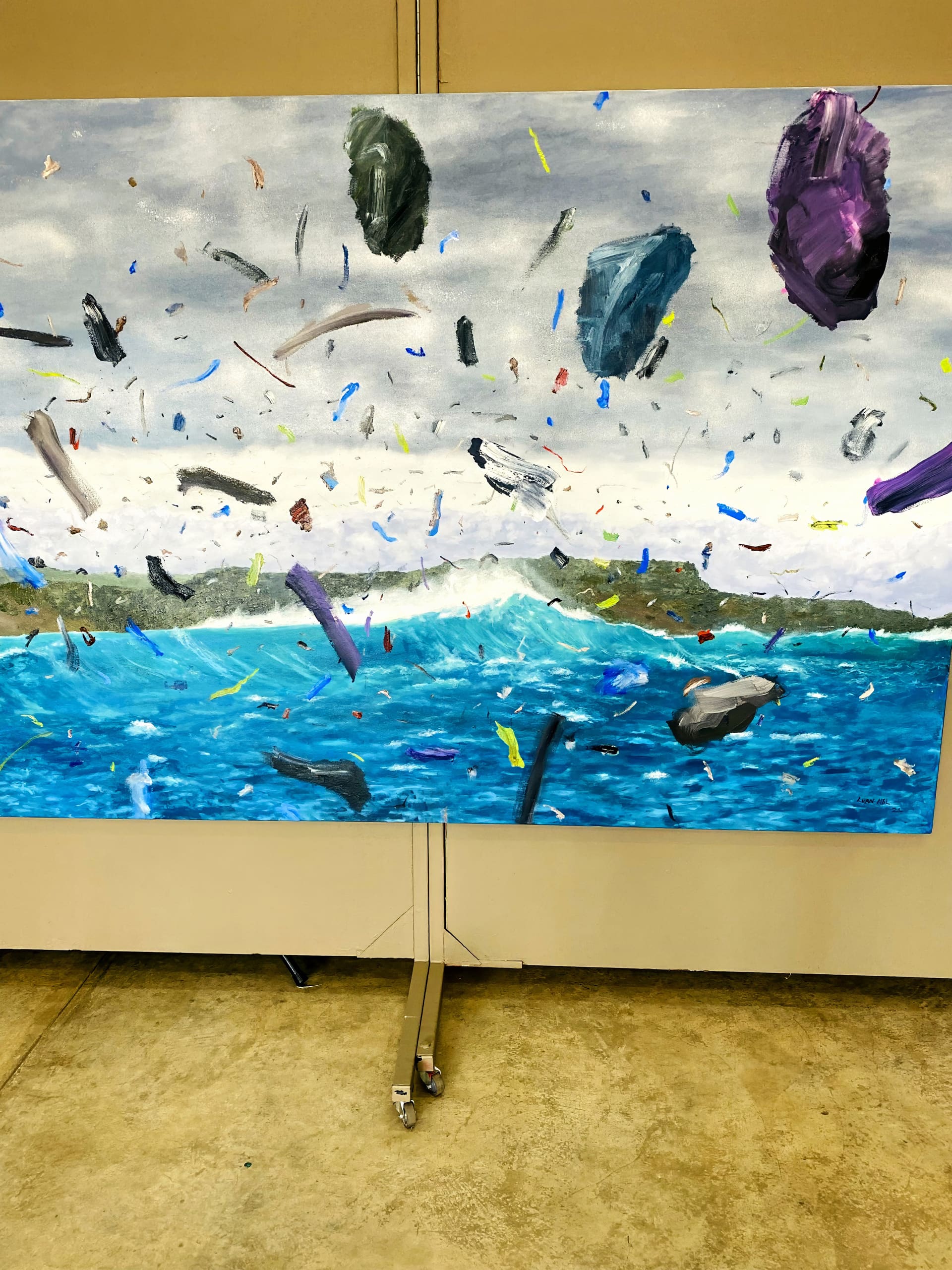 A colourful painting depicts a turbulent ocean under a cloudy sky, with scattered floating debris and fragments in mid-air, including variously sized rocks and objects, creating a sense of chaos and environmental disruption. The artwork is displayed on beige walls.