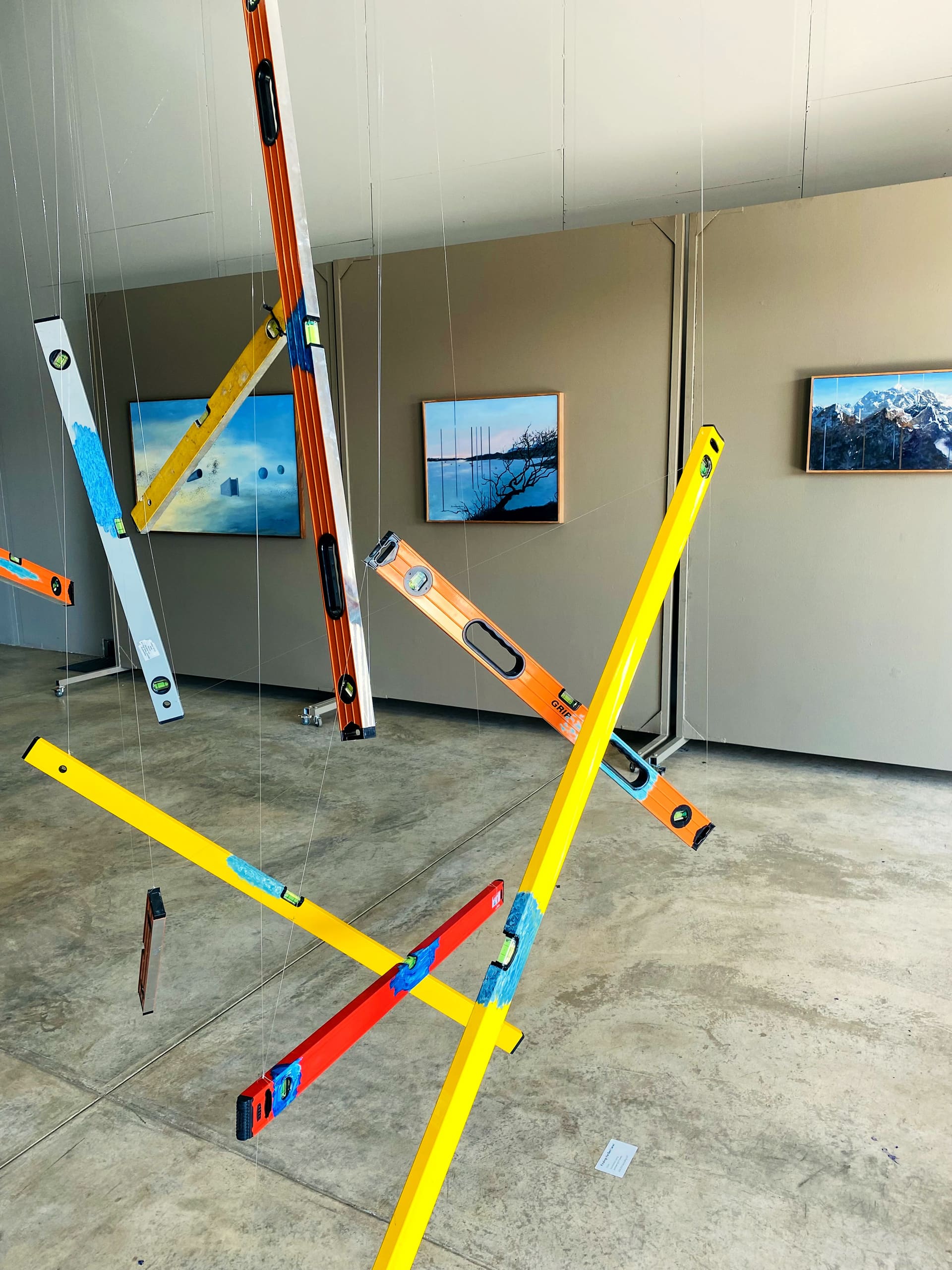 Several colourful spirit levels are suspended by thin strings in a modern gallery space with concrete floors. In the background, framed landscape photographs hang on beige walls, featuring water and nature scenes. The room is brightly lit with natural light.