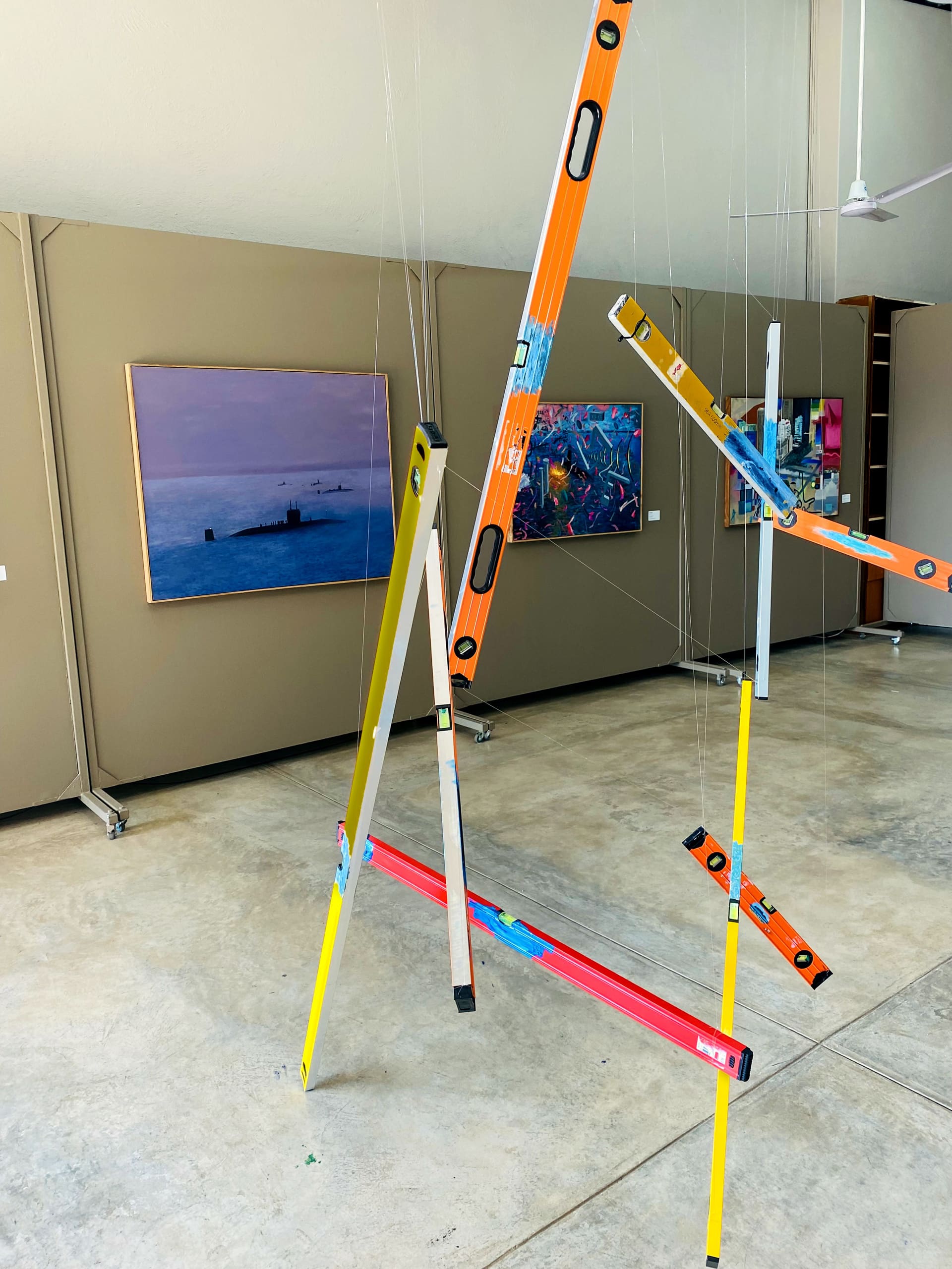 A sculpture made of colourful spirit levels suspended by thin wires stands on a concrete floor in an art gallery. Behind it, paintings hang on beige walls, including a seascape and abstract artworks. The space is bright and modern, with high ceilings.