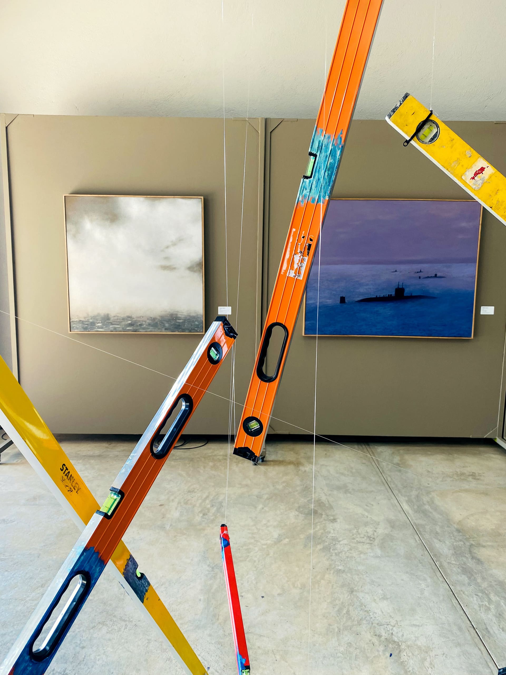 Several colourful spirit levels are suspended by thin strings in front of two paintings on a beige gallery wall. The left painting depicts a cloudy sky over water, while the right painting shows a blue-toned seascape with submarines.