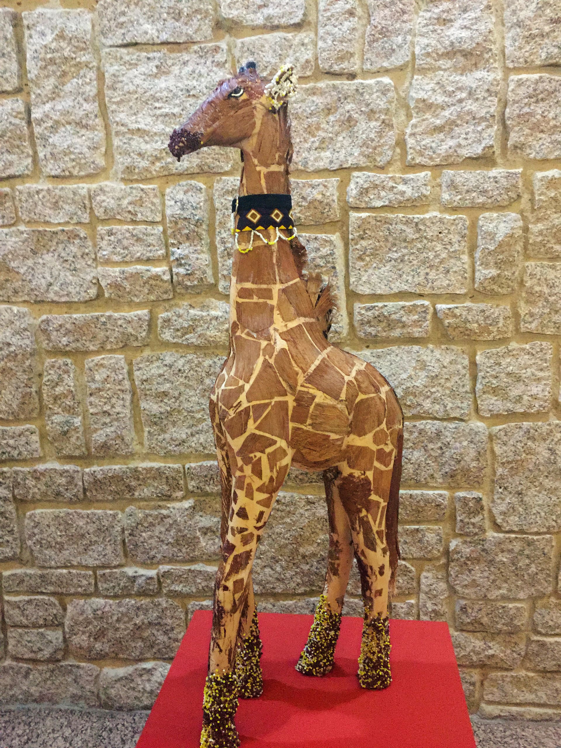 A hand-crafted giraffe sculpture with realistic brown and tan patterns stands on a red platform. It wears a dark blue necklace with a gold and yellow design, and its legs are decorated with golden beads. The background is a light stone wall.