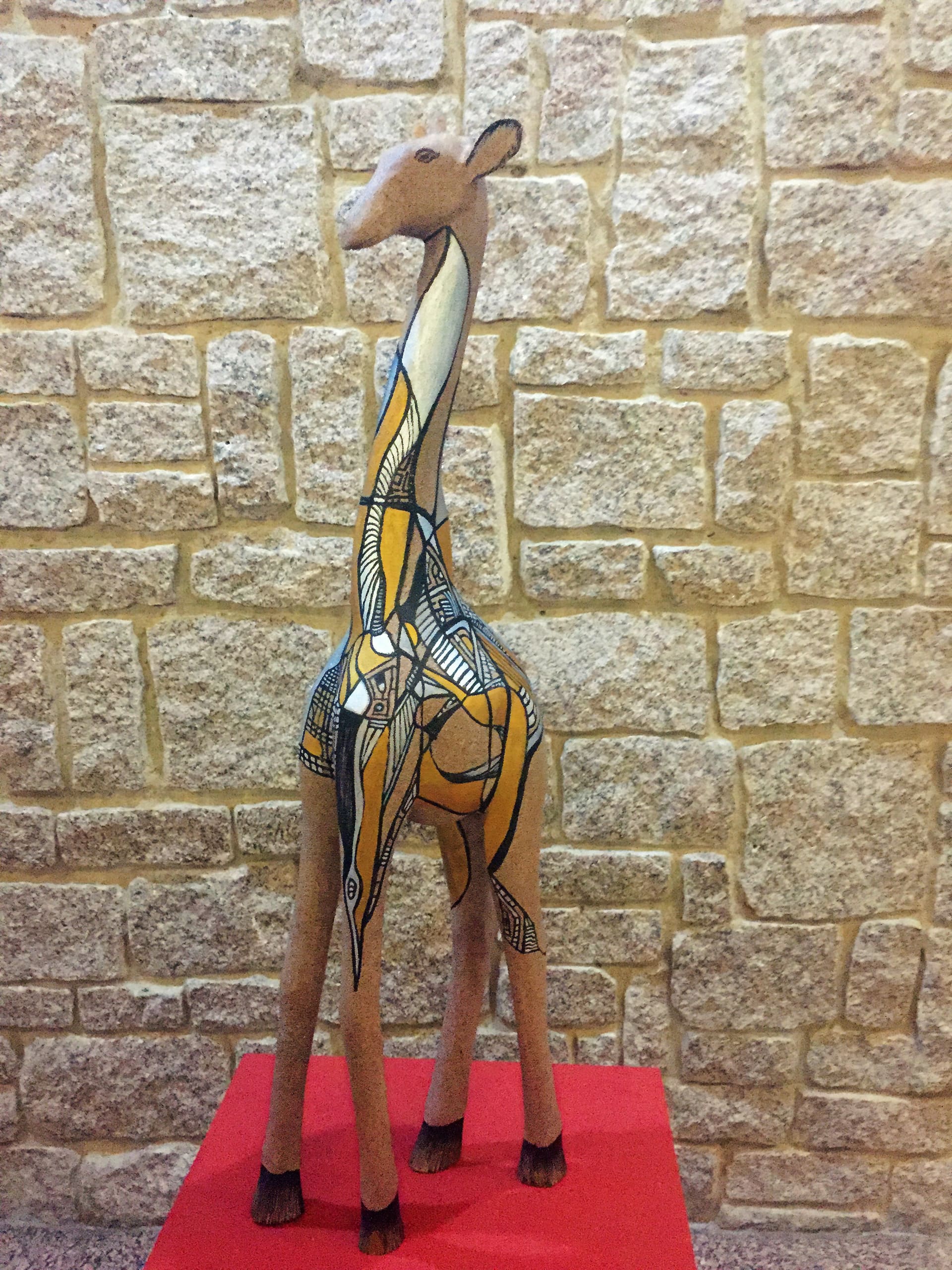 A stylised giraffe sculpture stands on a red platform against a beige stone wall. The giraffe features bold, abstract patterns in black, white, and yellow across its body, giving it a modern and artistic appearance.