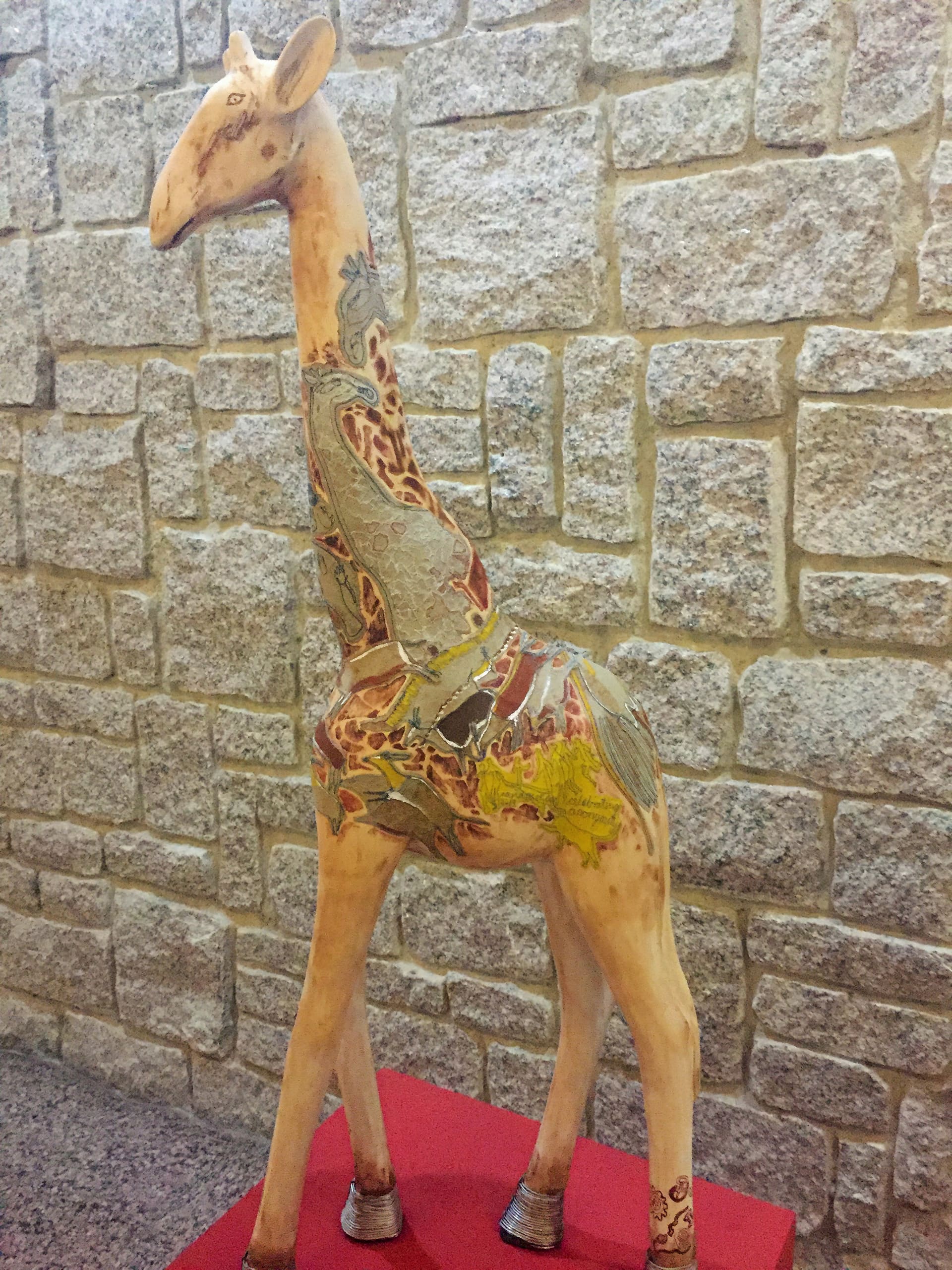 A painted giraffe sculpture with decorative patterns and earthy colours stands on a red platform in front of a textured stone wall. The patterns include leaves, animals, and abstract shapes on the giraffe’s body and neck.