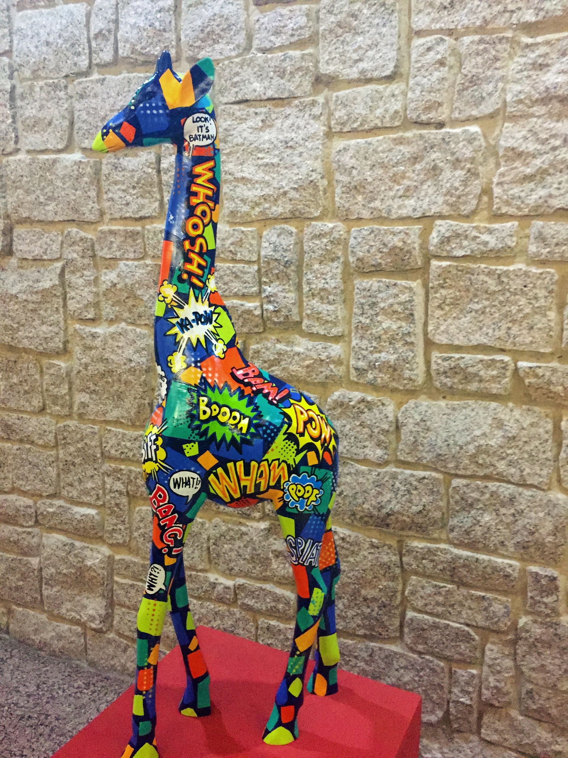A giraffe sculpture stands on a red platform against a stone wall. The giraffe is decorated with colourful comic book-style words and patterns, including 