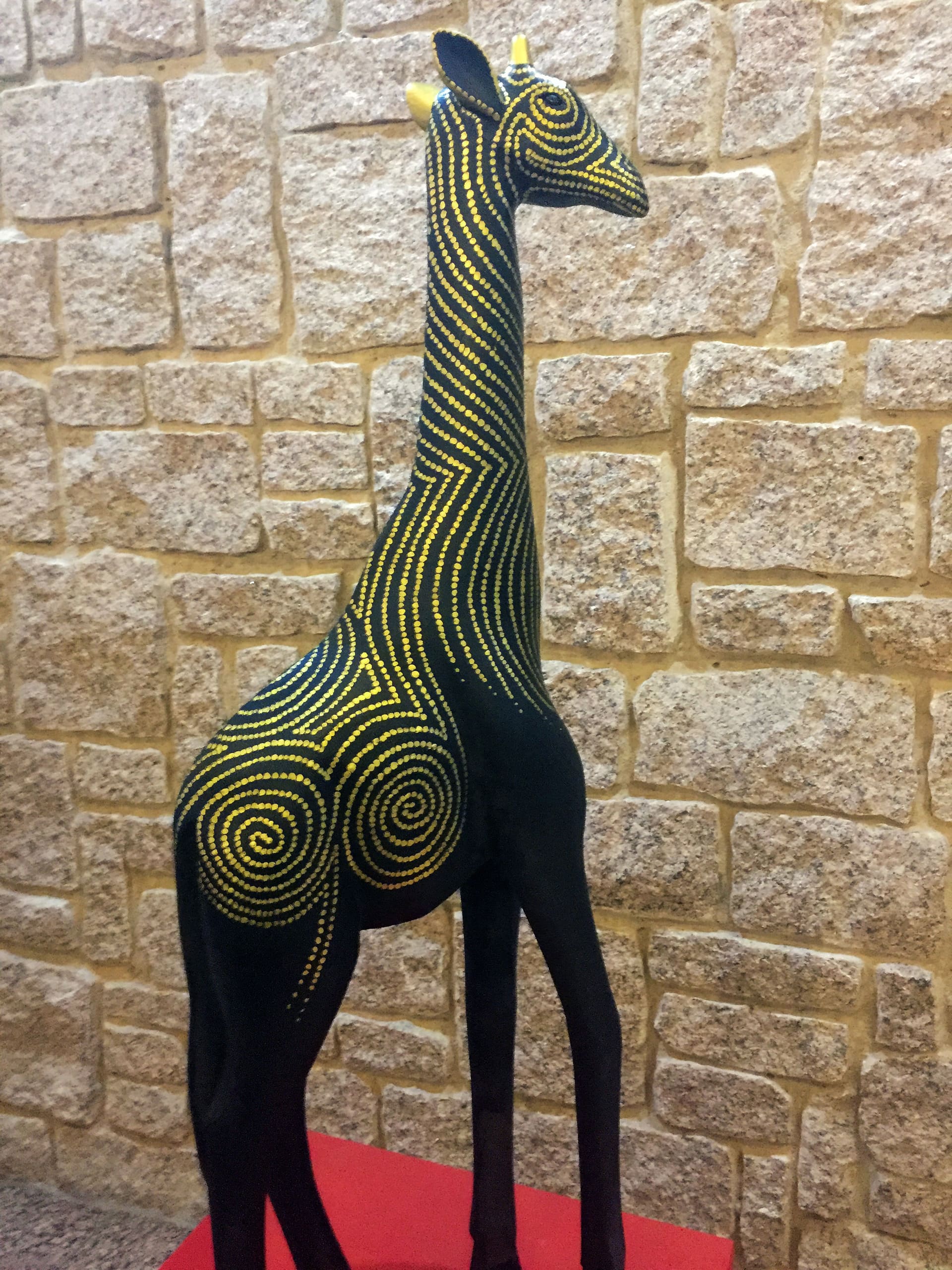 A black giraffe sculpture with intricate yellow dotted spiral patterns stands on a red surface in front of a textured beige stone wall. The artwork features elongated legs and neck, with stylised, geometric decoration covering its body.