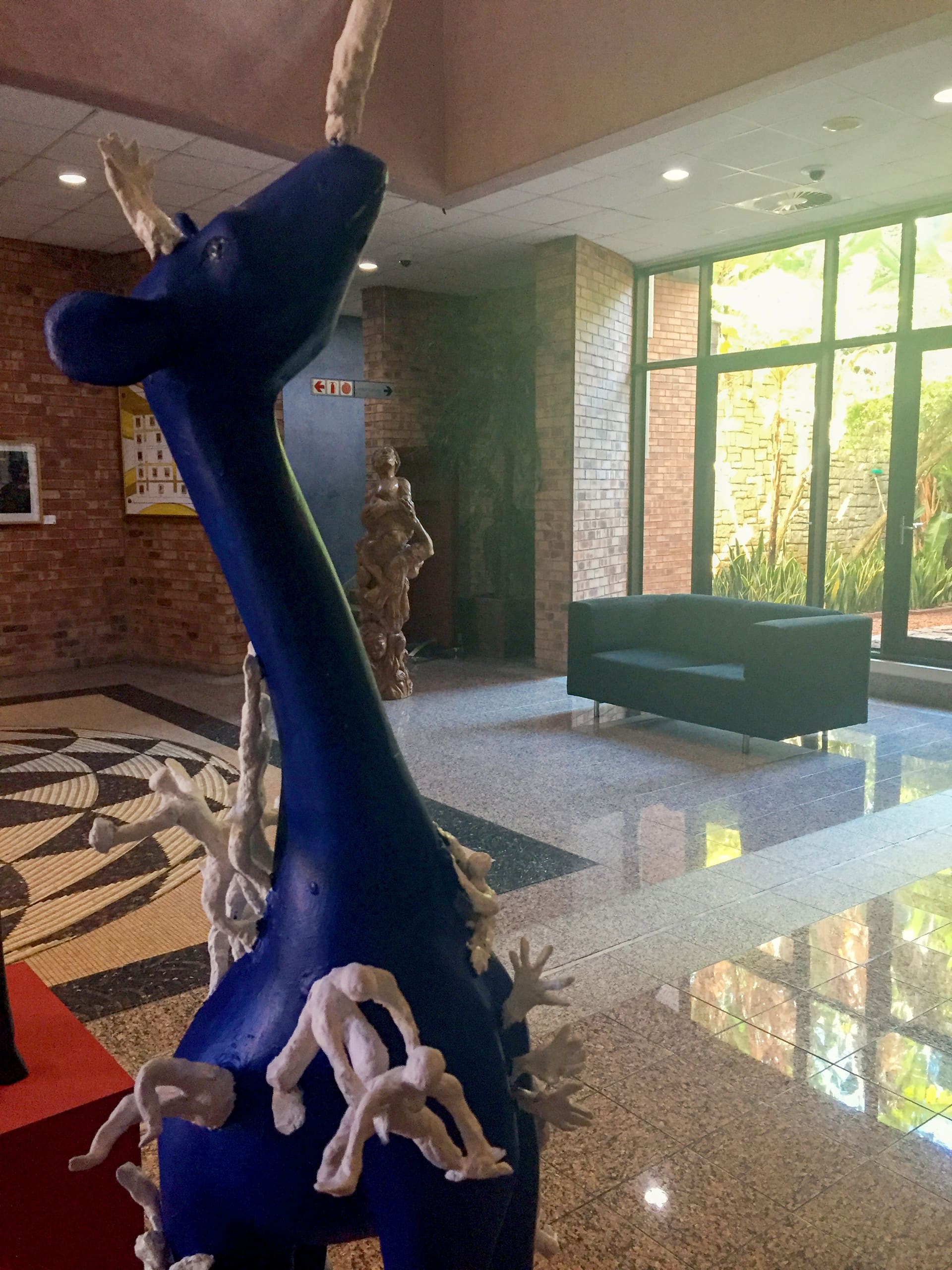 A blue abstract animal sculpture with a long neck and white, branch-like protrusions stands in a lobby with brick walls, large windows, a black sofa, and a mosaic tiled floor. Sunlight streams in, highlighting the indoor plants outside the window.
