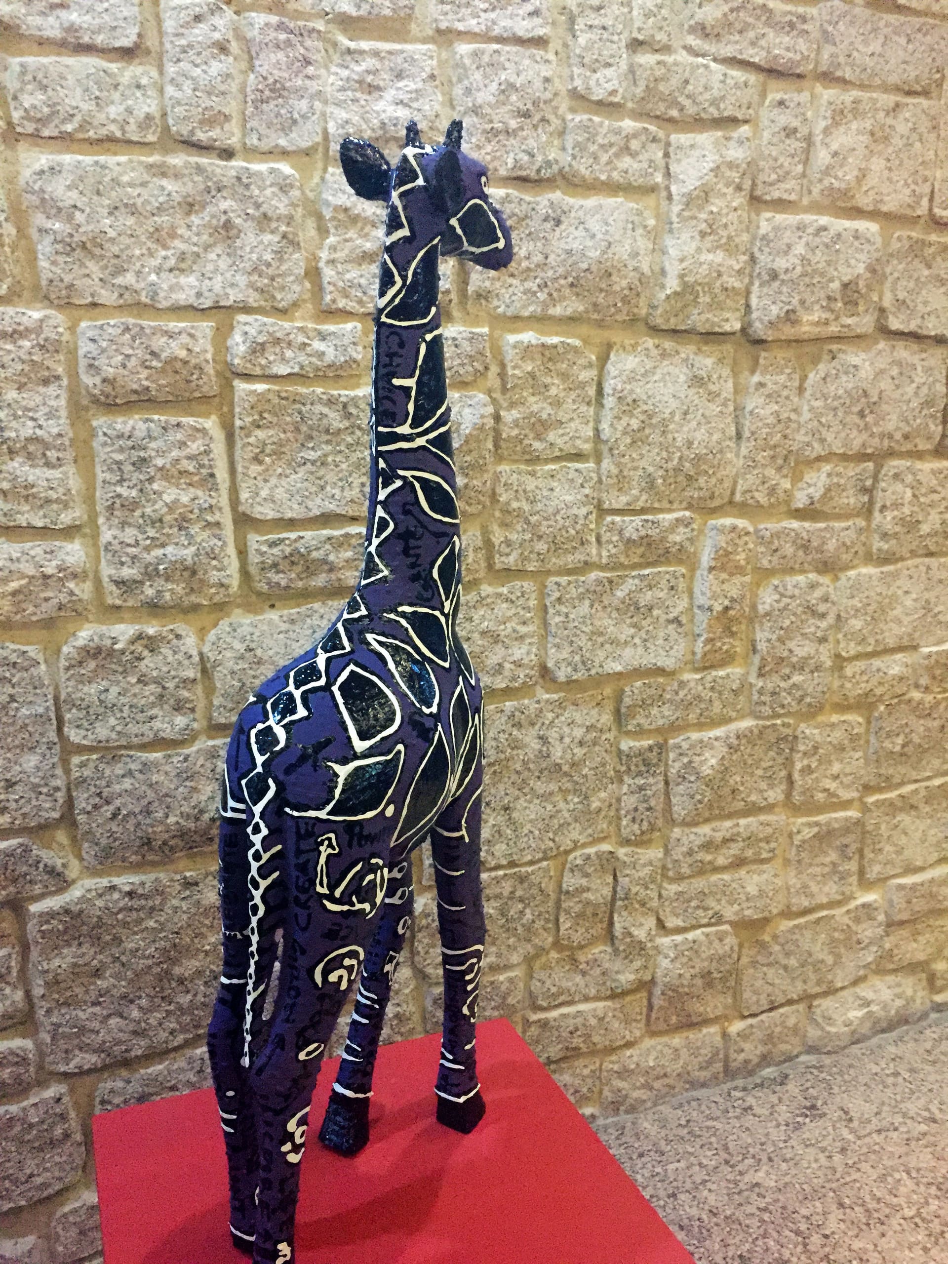 A tall, slender giraffe sculpture with abstract white and yellow designs stands on a red platform. The giraffe is dark purple and is set against a beige stone wall with large, rectangular blocks.