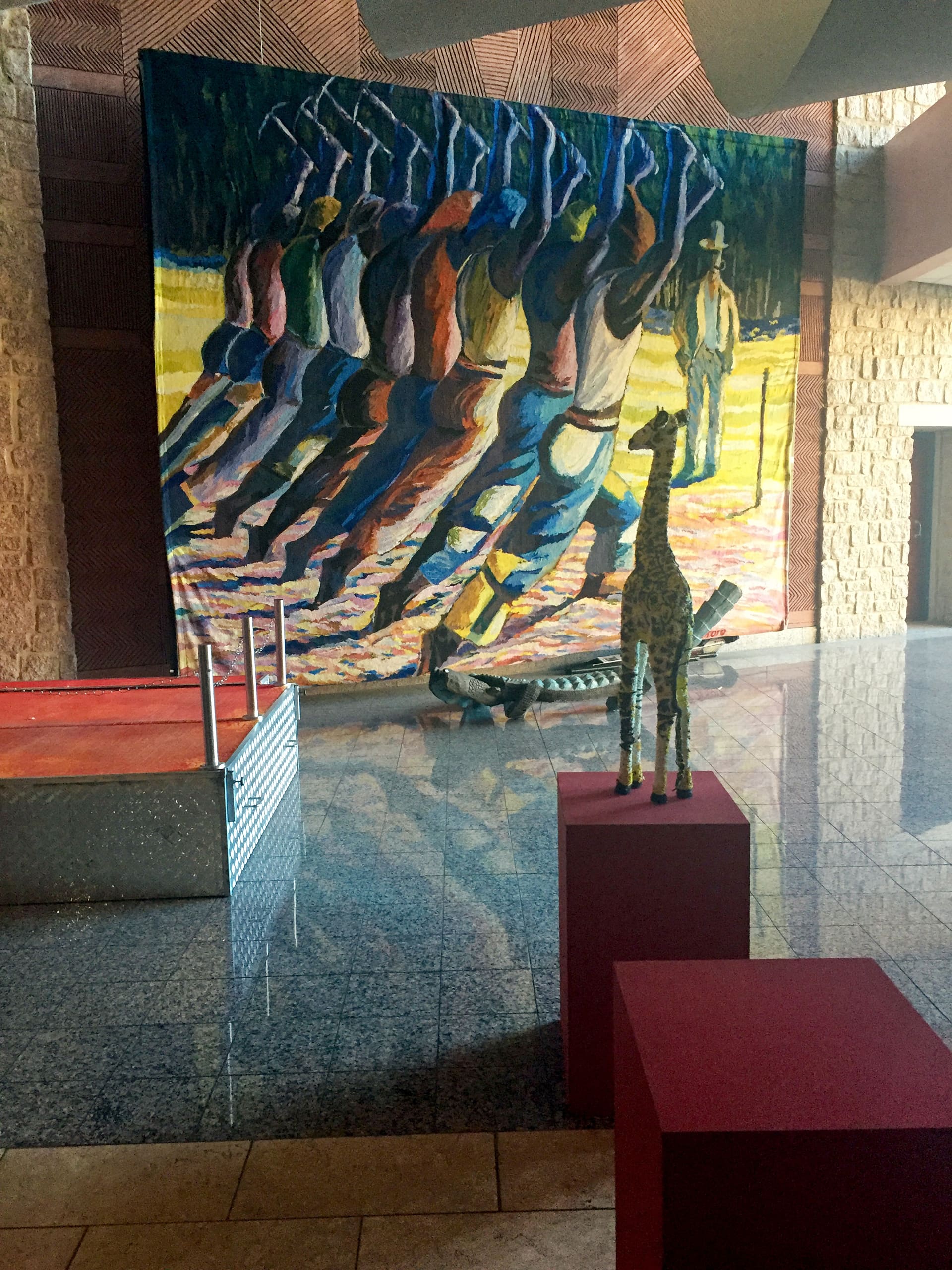 A brightly lit lobby features a colourful mural of people raising tools, a giraffe sculpture on a red plinth, and reflections on the shiny marble floor. The walls are textured stone, and light streams in from the right side.
