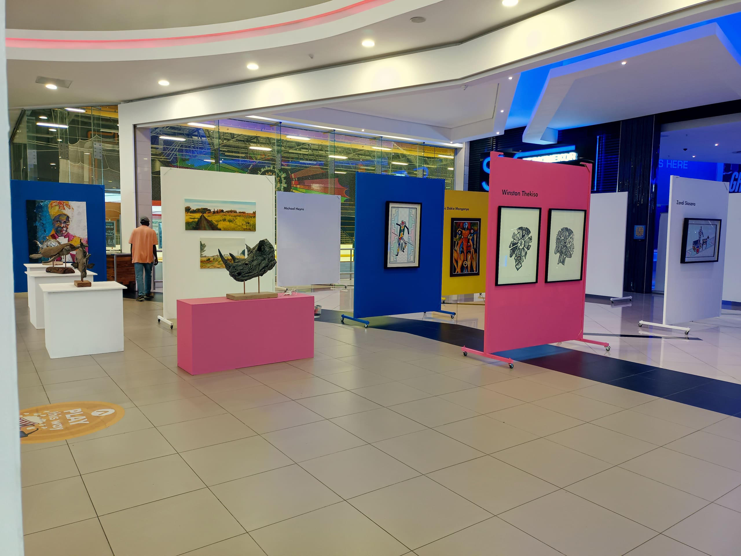 An art exhibition in a modern, brightly lit indoor space features colourful display boards with framed artworks, sculptures on white stands, and a few visitors viewing the exhibits. The floor is tiled, and signage and lighting add a contemporary feel.