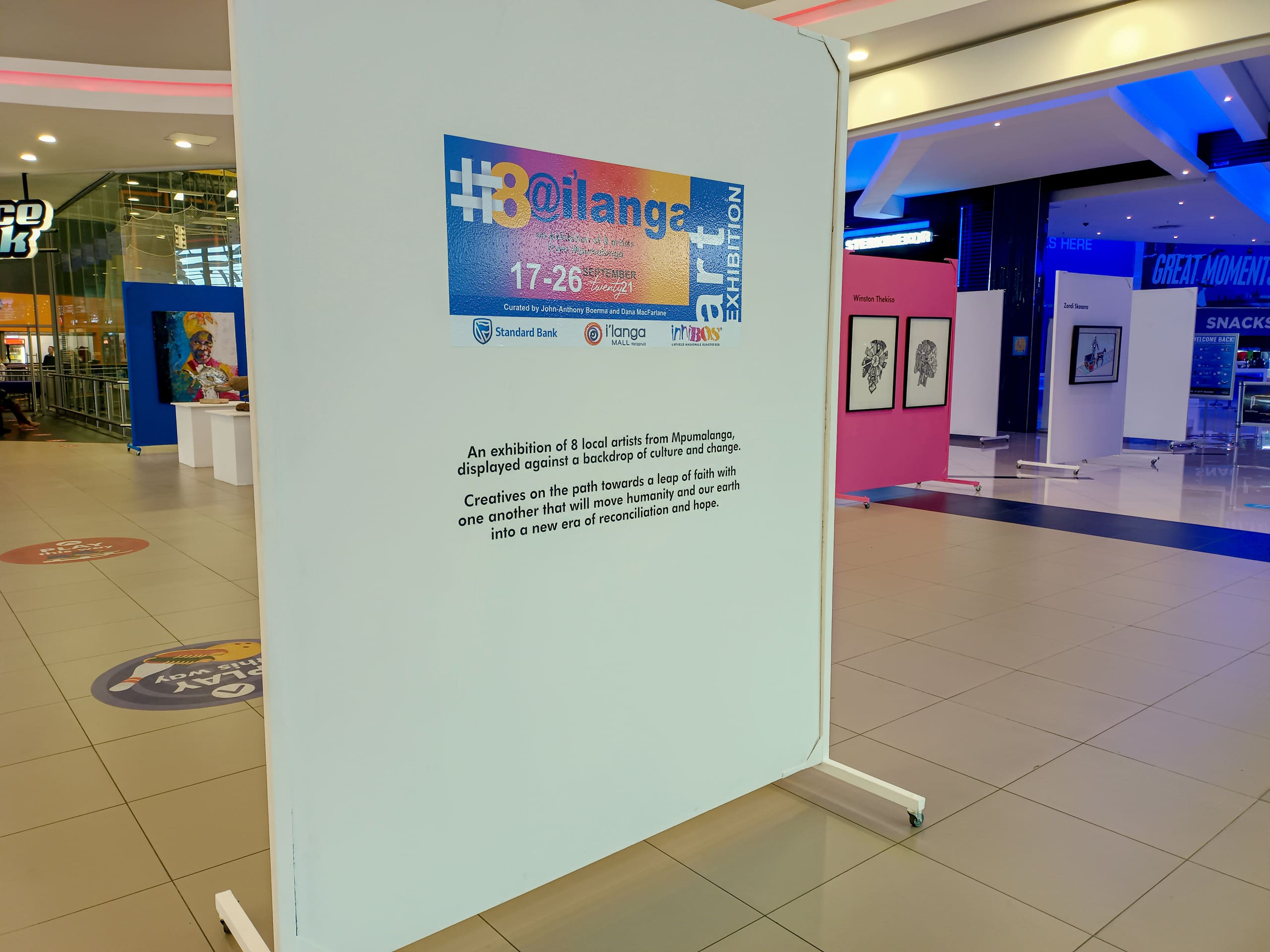 A white exhibition panel in a shopping centre describes the “#30@ilanga” art exhibition, with logos at the top. The space is open, with framed artworks on easels in the background and shopfronts visible, including a sign for GREAT MOVIES.
