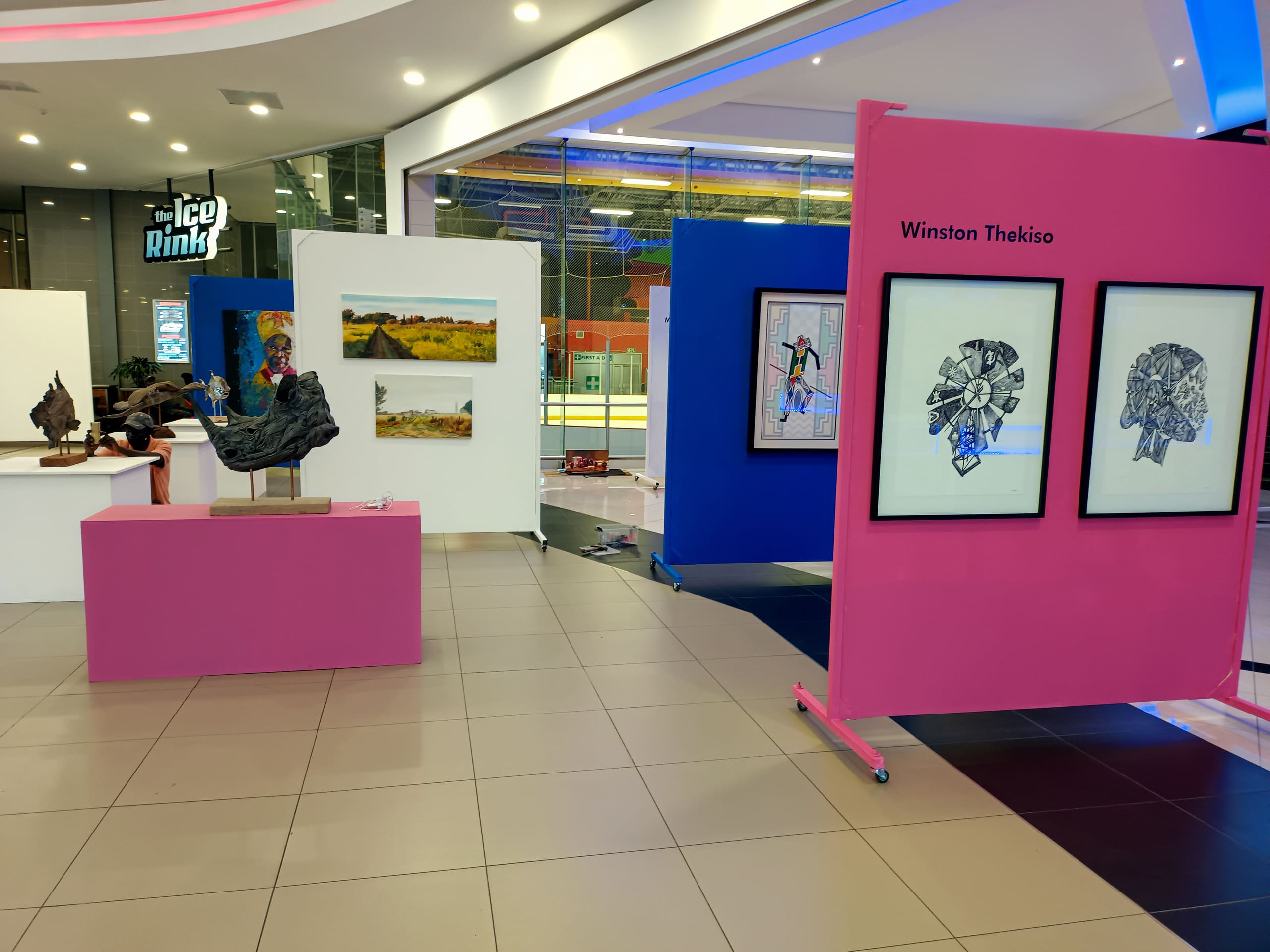 An indoor art exhibition with colourful panels displays paintings, sculptures, and framed art. A pink panel features Winston Thekiso’s name and two abstract works. In the background, there’s an “Ice Rink” sign and glass walls revealing a skating area.
