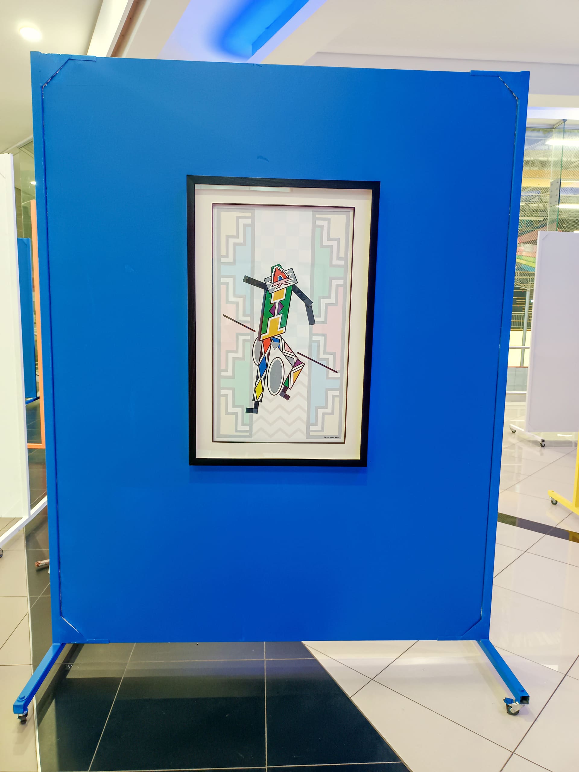 A framed abstract artwork featuring geometric shapes and bold colours hangs on a large blue display board with wheels. The background includes tiled flooring and glass walls, suggesting an indoor exhibition space.