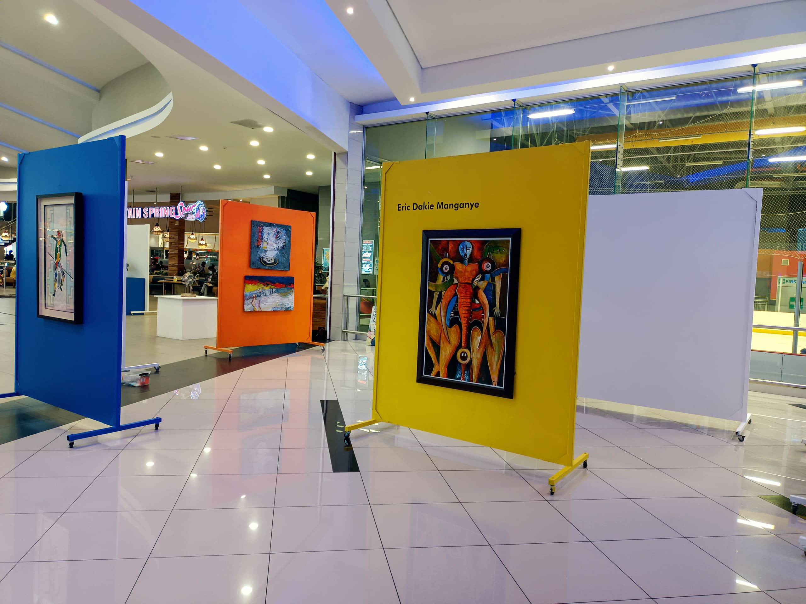 An indoor art display shows three colourful partition walls—orange, yellow, and white—each featuring framed artwork. The yellow wall displays a bold, abstract painting by Eric Dalie Manganye. The setting is a modern shopping centre with shiny tiled floors.