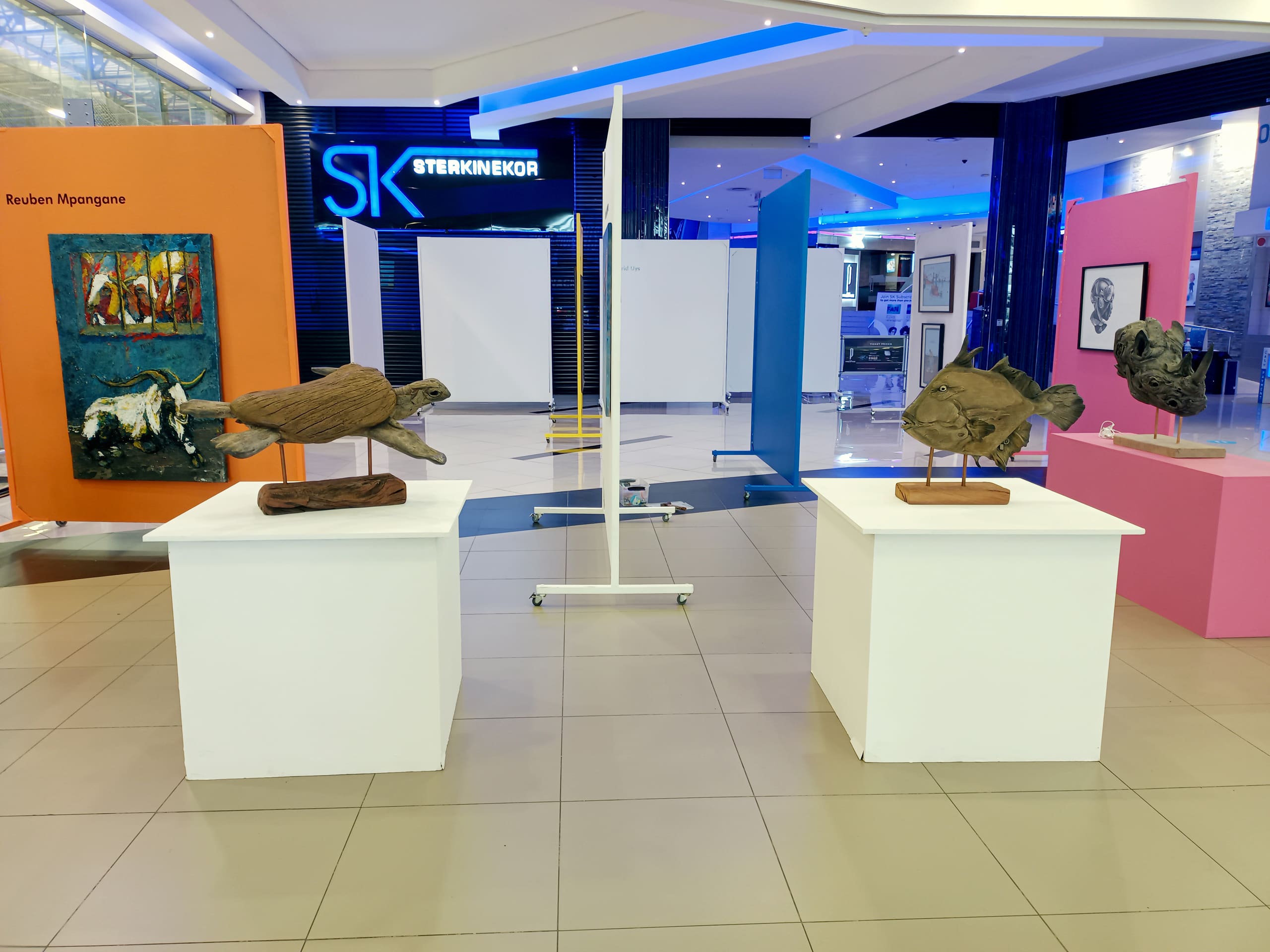 An art gallery with sculptures on white plinths, colourful walls displaying paintings, and a Ster-Kinekor cinema sign in the background. The space is brightly lit with blue and white lighting accents.