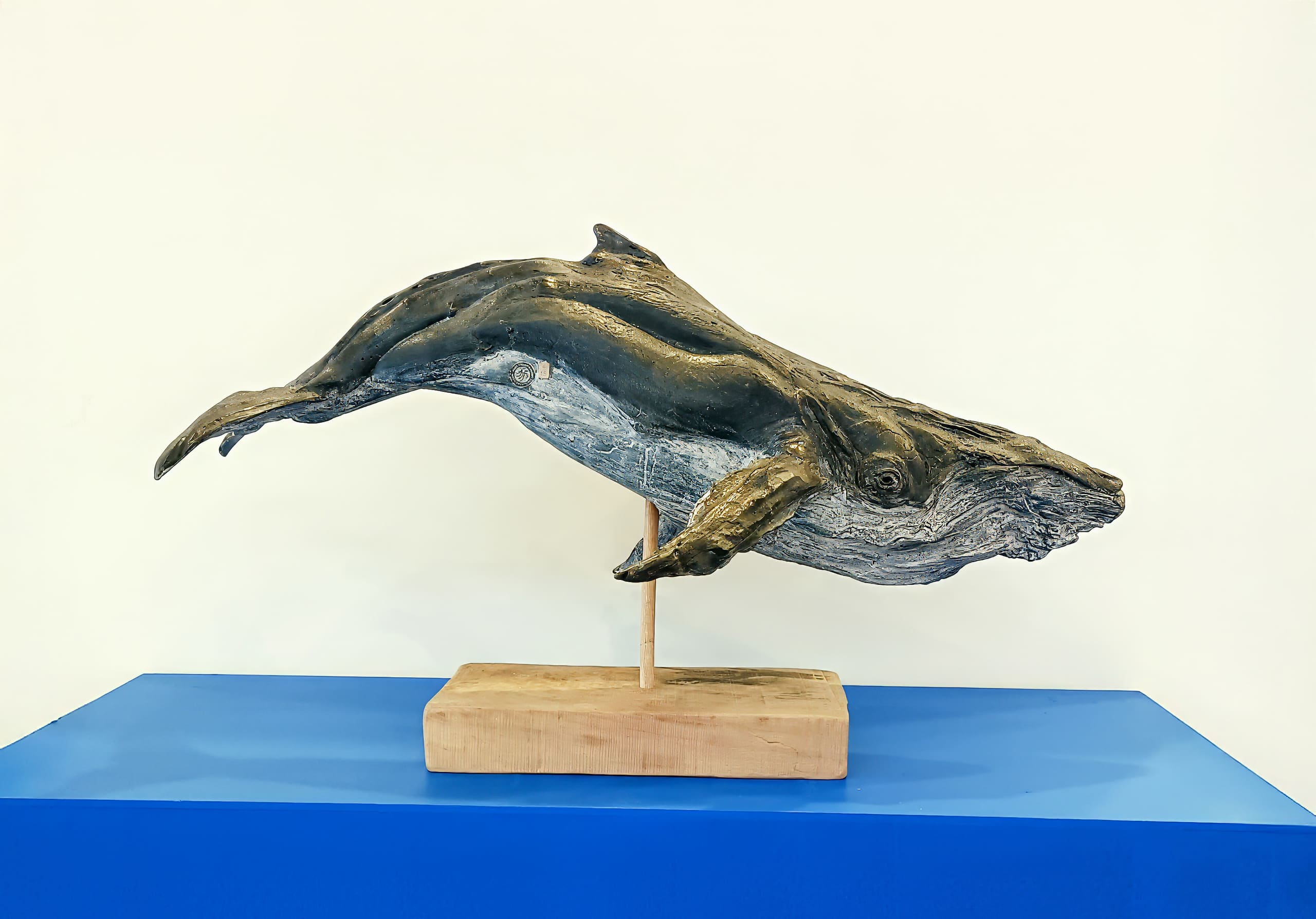 A realistic whale sculpture made of textured material, painted dark grey with lighter accents, is mounted on a wooden base. The sculpture is displayed on a bright blue surface against a plain white background.