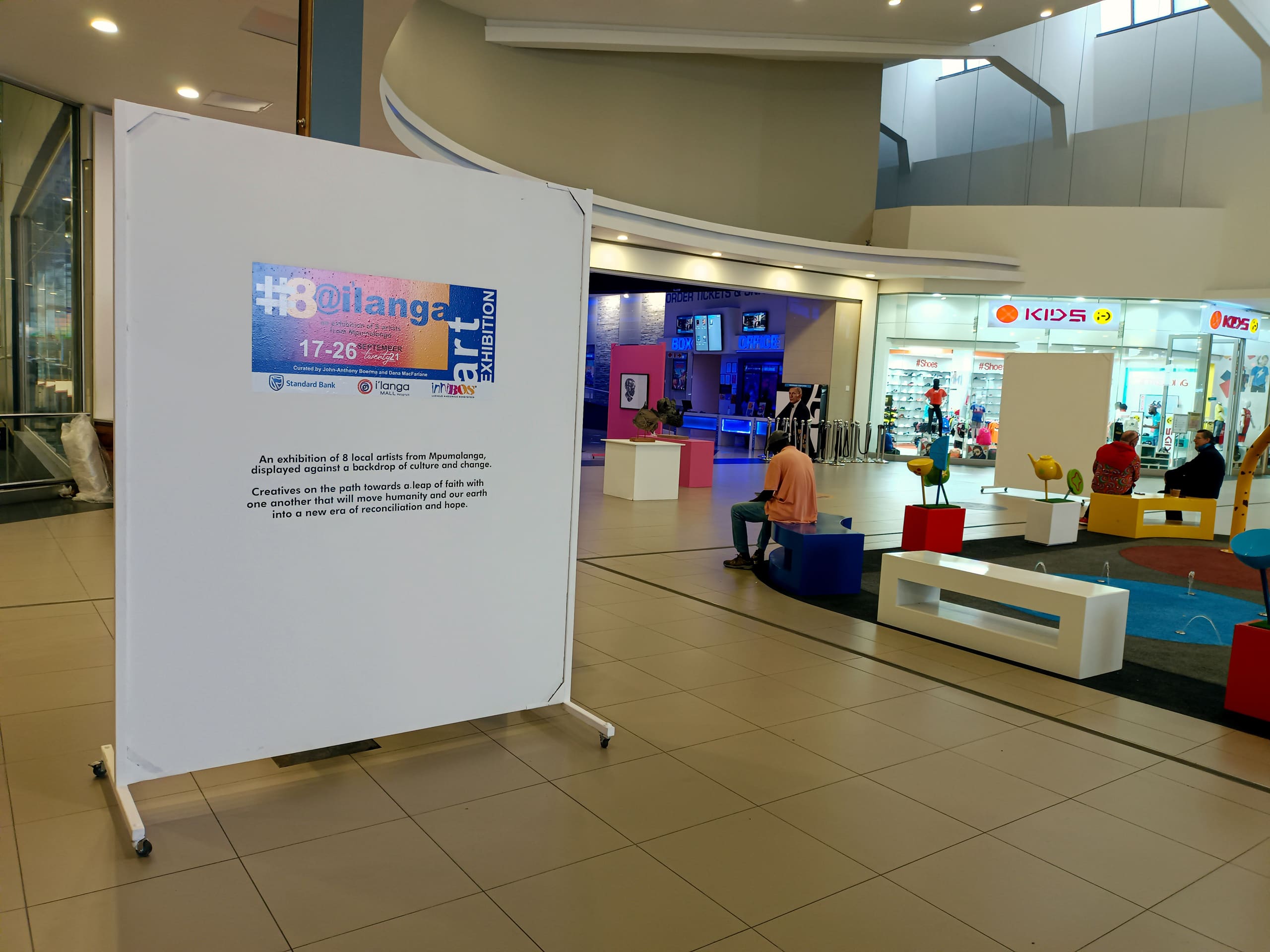 A white exhibition board stands in a shopping centre with a poster for “#OilangArt Exhibition 17–26,” surrounded by shops, colourful seating, and a children’s play area; people are casually seated and walking in the open, well-lit space.