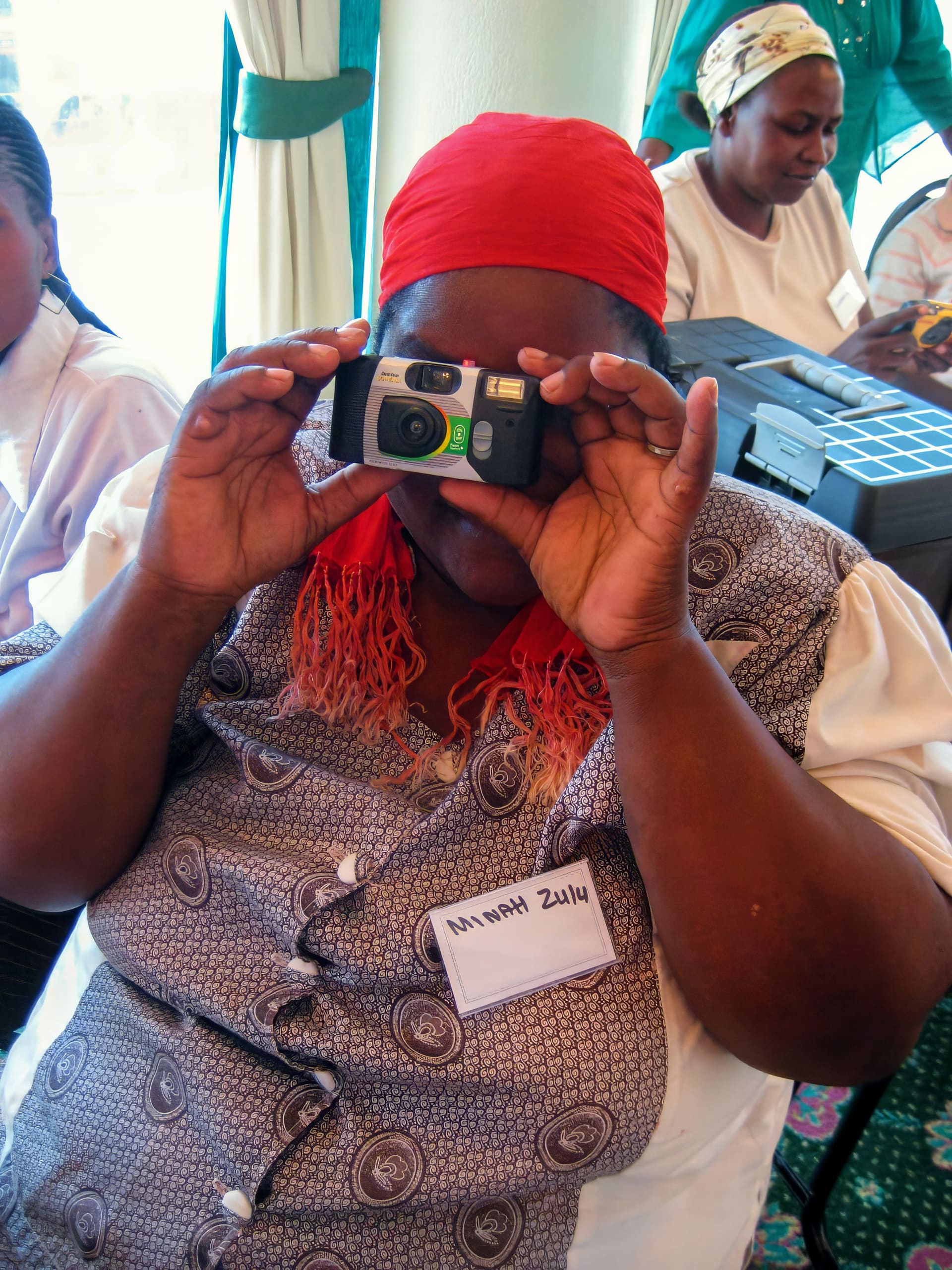 A woman wearing a red headscarf and patterned waistcoat holds a disposable camera up to her face. She has a name tag reading 