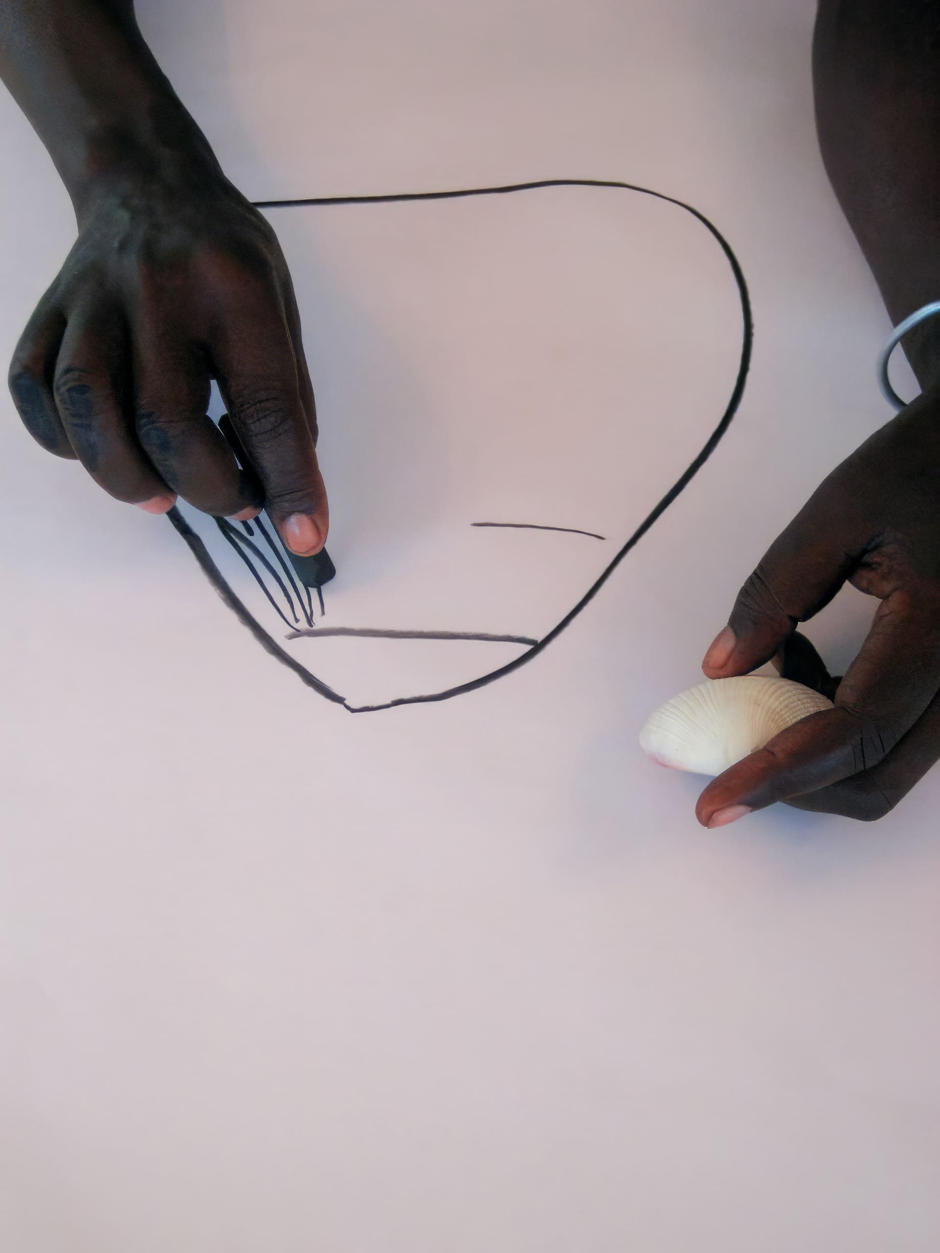 Two dark-skinned hands work on a white surface. One hand holds a black marker, drawing lines inside a large outlined shape, while the other hand holds a white seashell. The shape resembles a face or mask, and the background is plain.