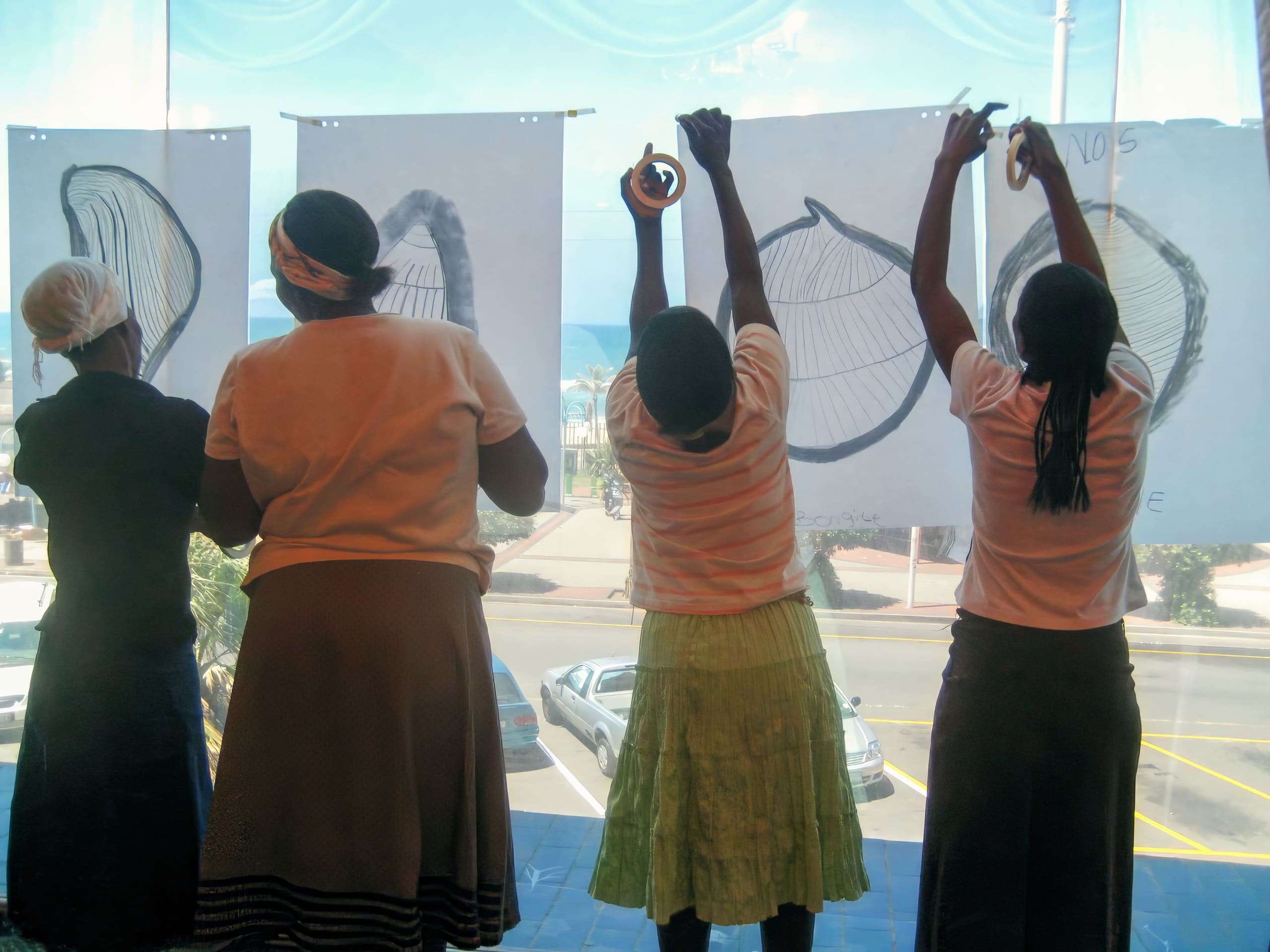 Four people stand indoors facing large windows, hanging up drawings of seashells on sheets of white paper. Sunlight streams in, casting their silhouettes. Outside, cars, palm trees, and the sea are visible through the glass.