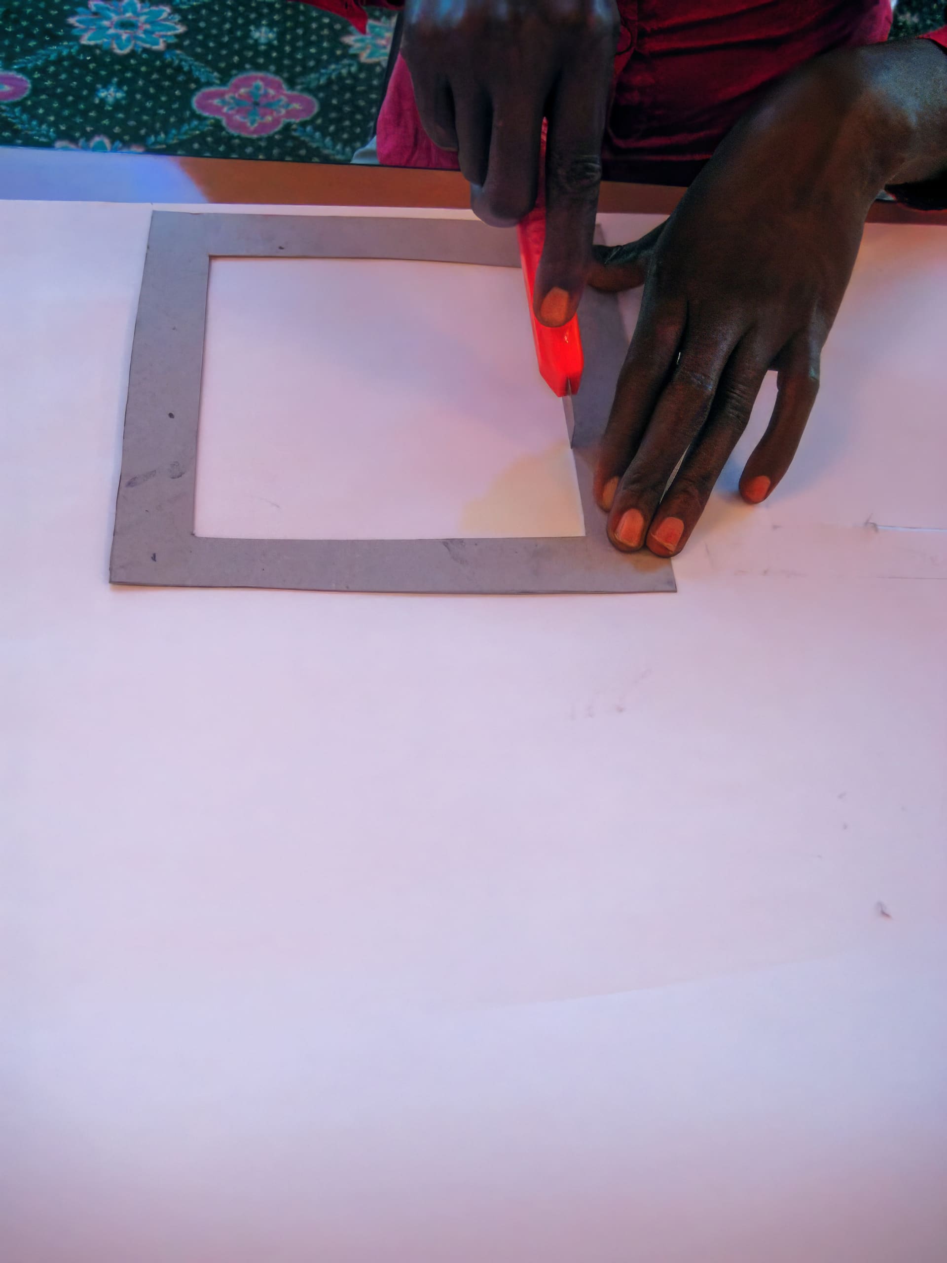 A person holds a cardboard frame on white paper with one hand whilst cutting along its edge using a red utility knife. Only the hands and part of the frame are visible. The background includes a patterned green surface.