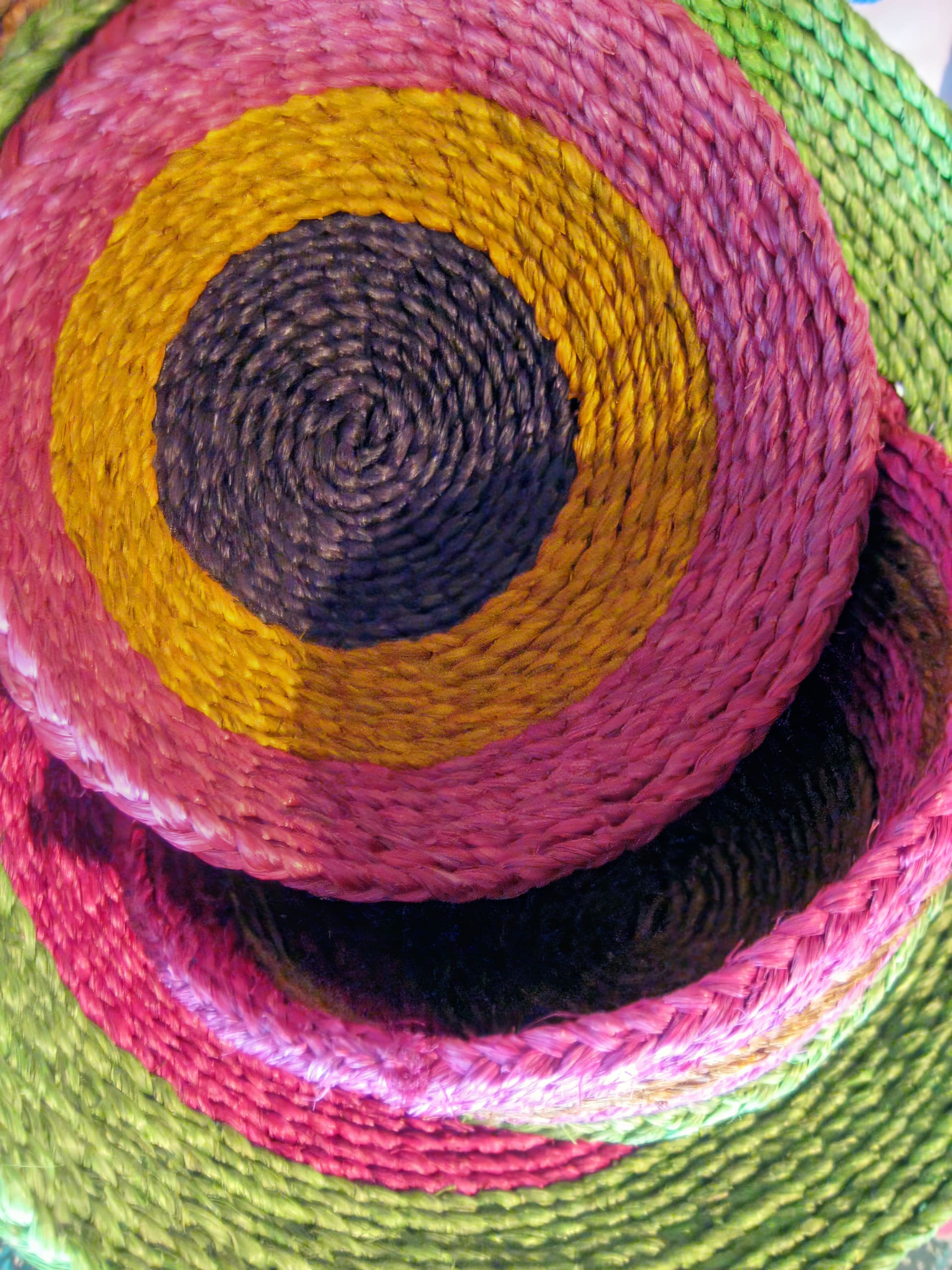 Close-up of two woven, circular baskets stacked together. The top basket has concentric rings in black, yellow, and pink, whilst the bottom one features rings in green and pink. The texture of the tightly woven fibres is clearly visible.