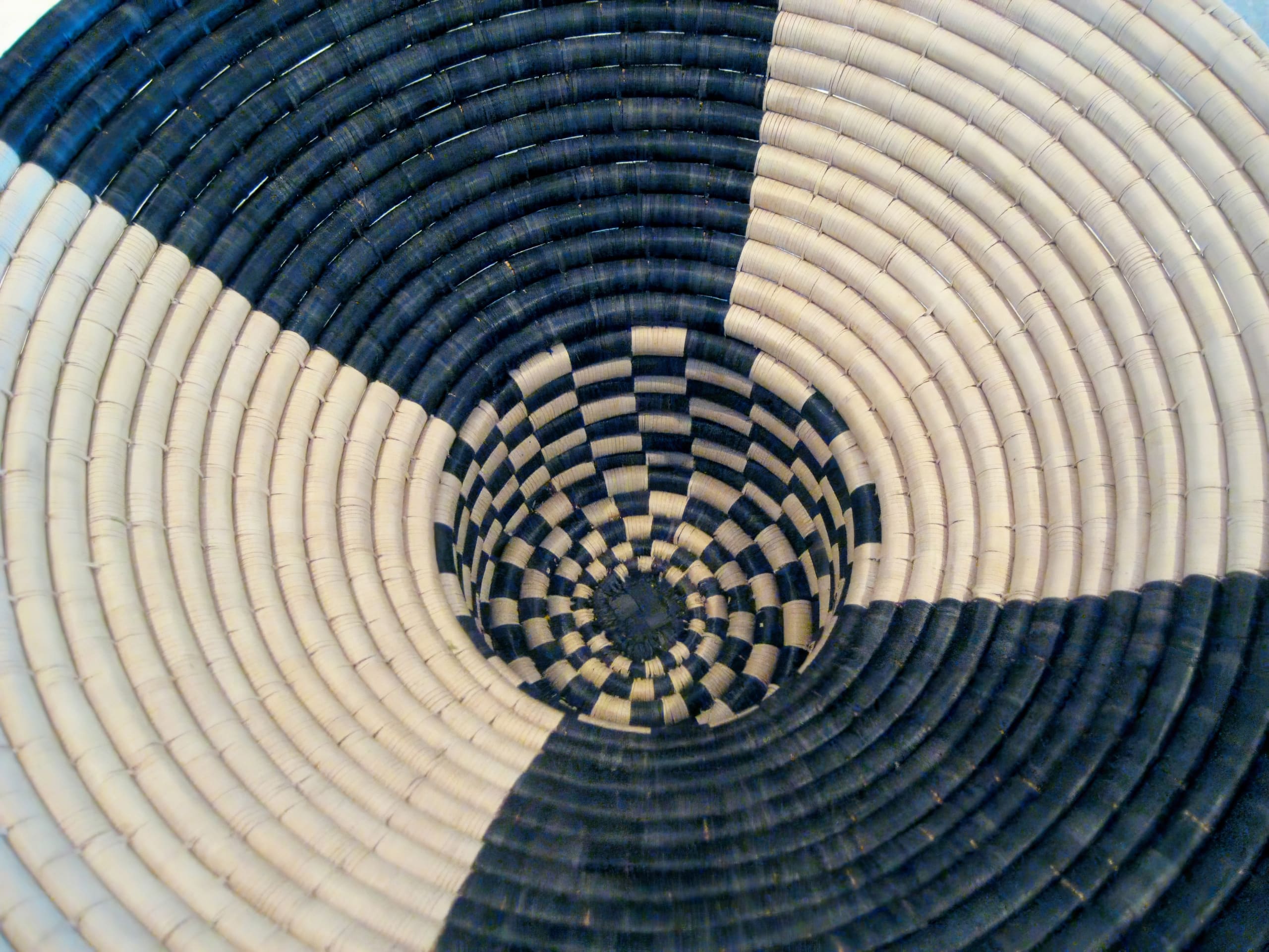 A woven basket with a spiral pattern in alternating black and natural beige sections, forming a geometric, hypnotic design that radiates from the centre outward; the fibre texture is visible, showing intricate craftsmanship.