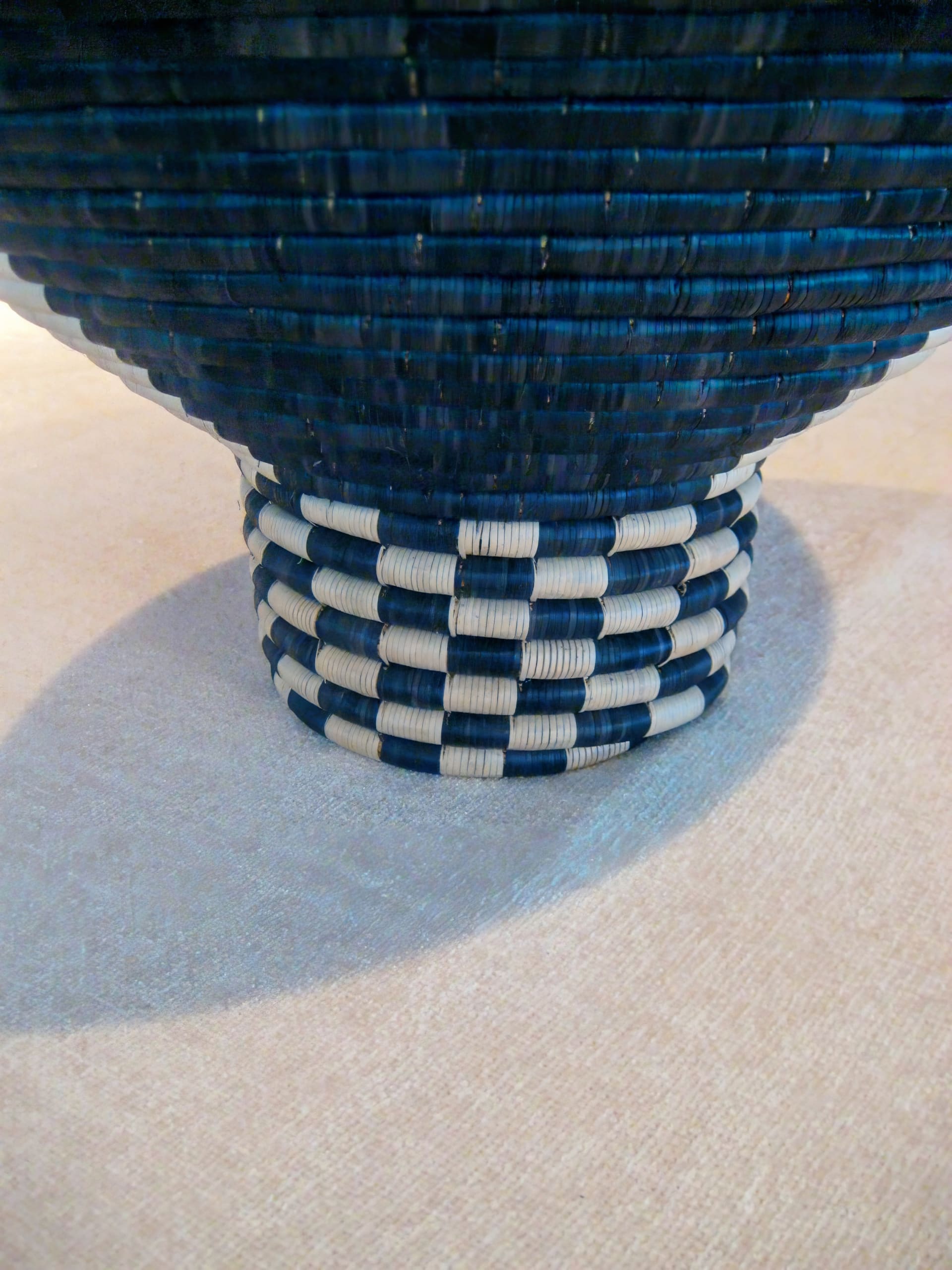 A close-up of the base of a woven basket with dark blue and white alternating bands in a chequered pattern. The surface underneath is a light beige fabric, and the shadow of the basket is visible on the left side.