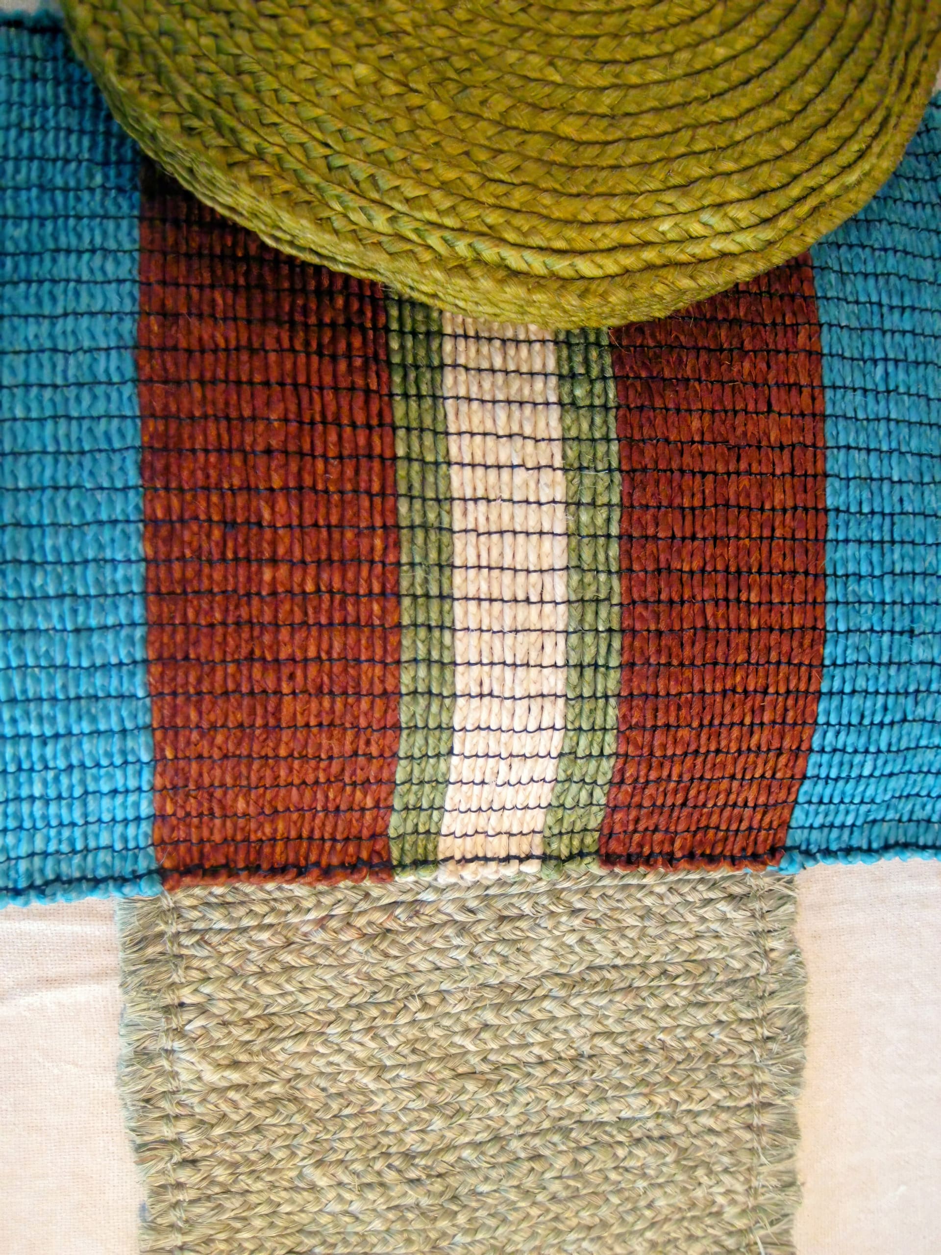 A close-up of a woven textile featuring vertical stripes in blue, rust red, green, and cream, with a circular woven green element at the top and a rectangular section of green textured weaving near the bottom. The patterns and textures are clearly visible.
