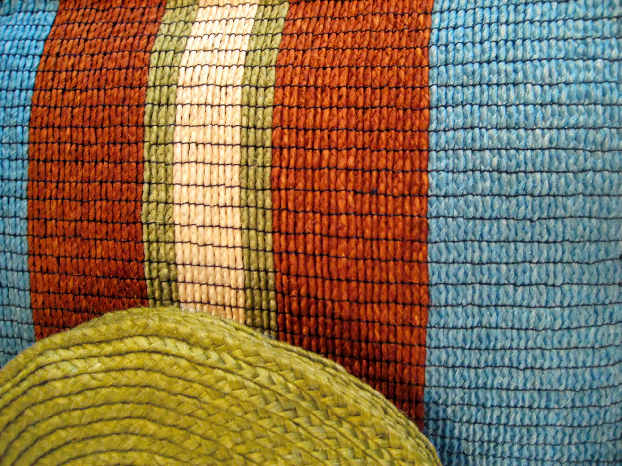 Close-up of a woven textile featuring blue and reddish-brown vertical stripes, with a thin cream stripe and a wider olive green stripe. In the lower left corner, part of a round, green woven basket or mat is visible, emphasising texture and colour contrast.