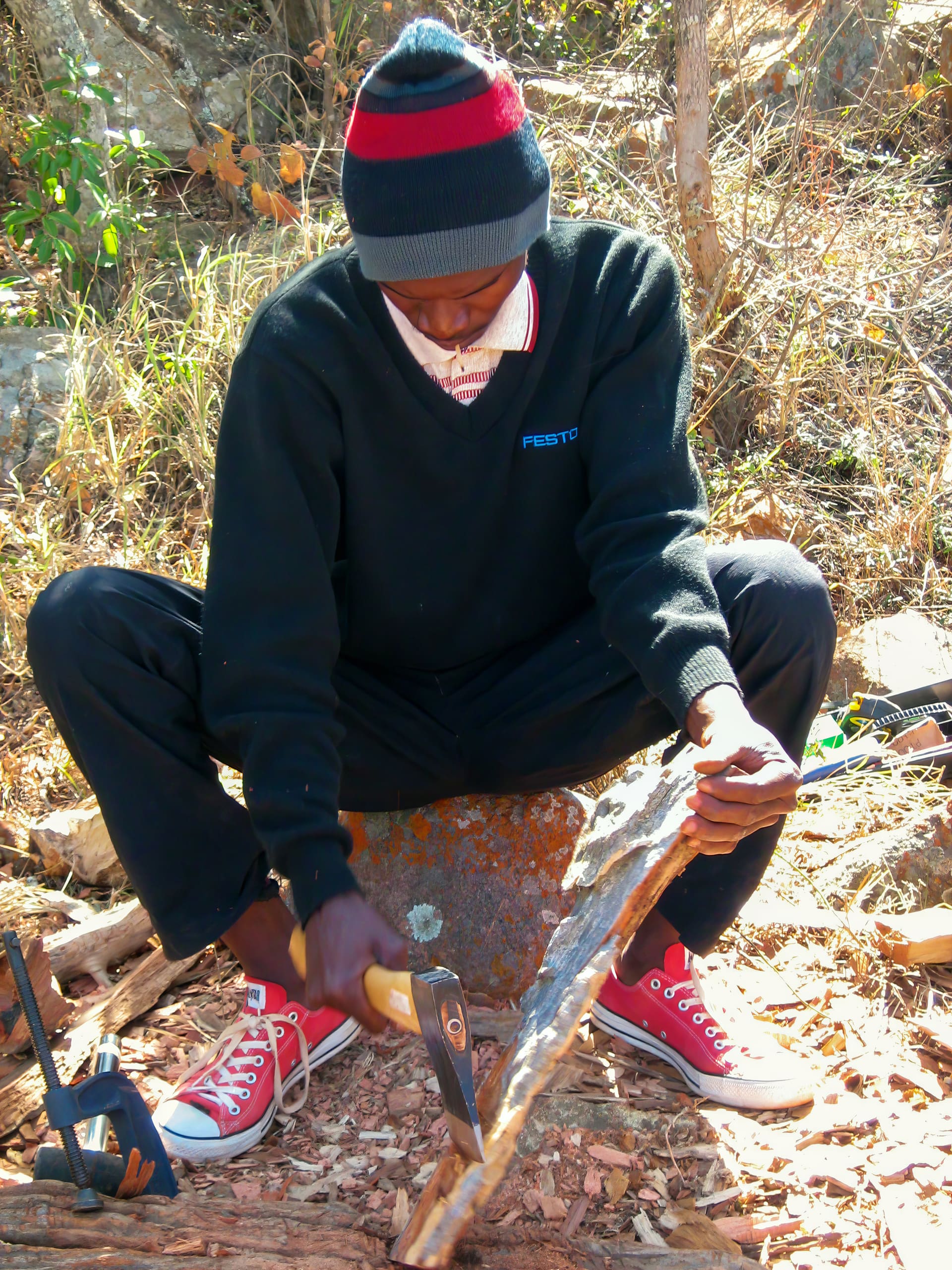 A person wearing a black jumper, red and black hat, red trainers, and a white mask saws a piece of wood while sitting outdoors on a rock surrounded by dry grass and leaves. The sunlight highlights the earthy, natural setting.