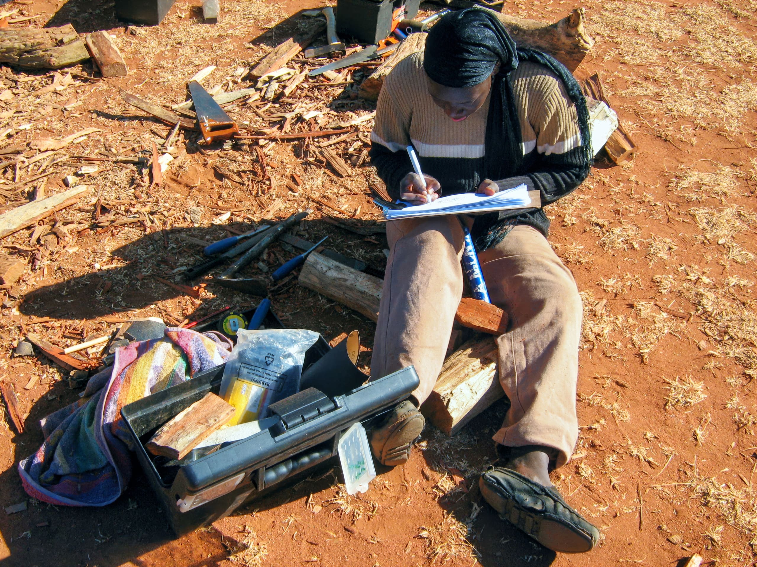 A person sits on reddish earth beside a toolbox, writing in a notebook. They wear a striped jumper, tan trousers, and a headscarf. Around them are scattered wooden sticks and tools, suggesting they’re working outdoors, possibly conducting research or fieldwork.