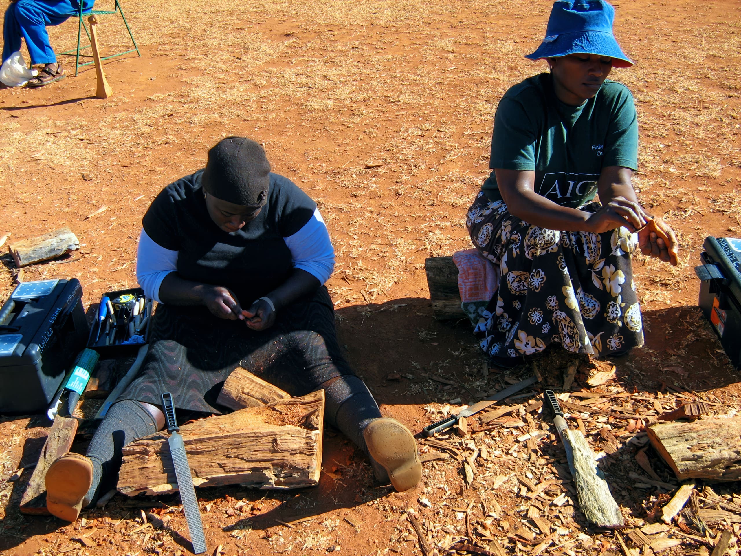 Two women sit on the ground outdoors on reddish earth, working with tools and wood. One woman is filing or carving a piece of wood, while the other holds a tool. Logs, toolboxes, and wood shavings are scattered around them. Both wear hats and long skirts.