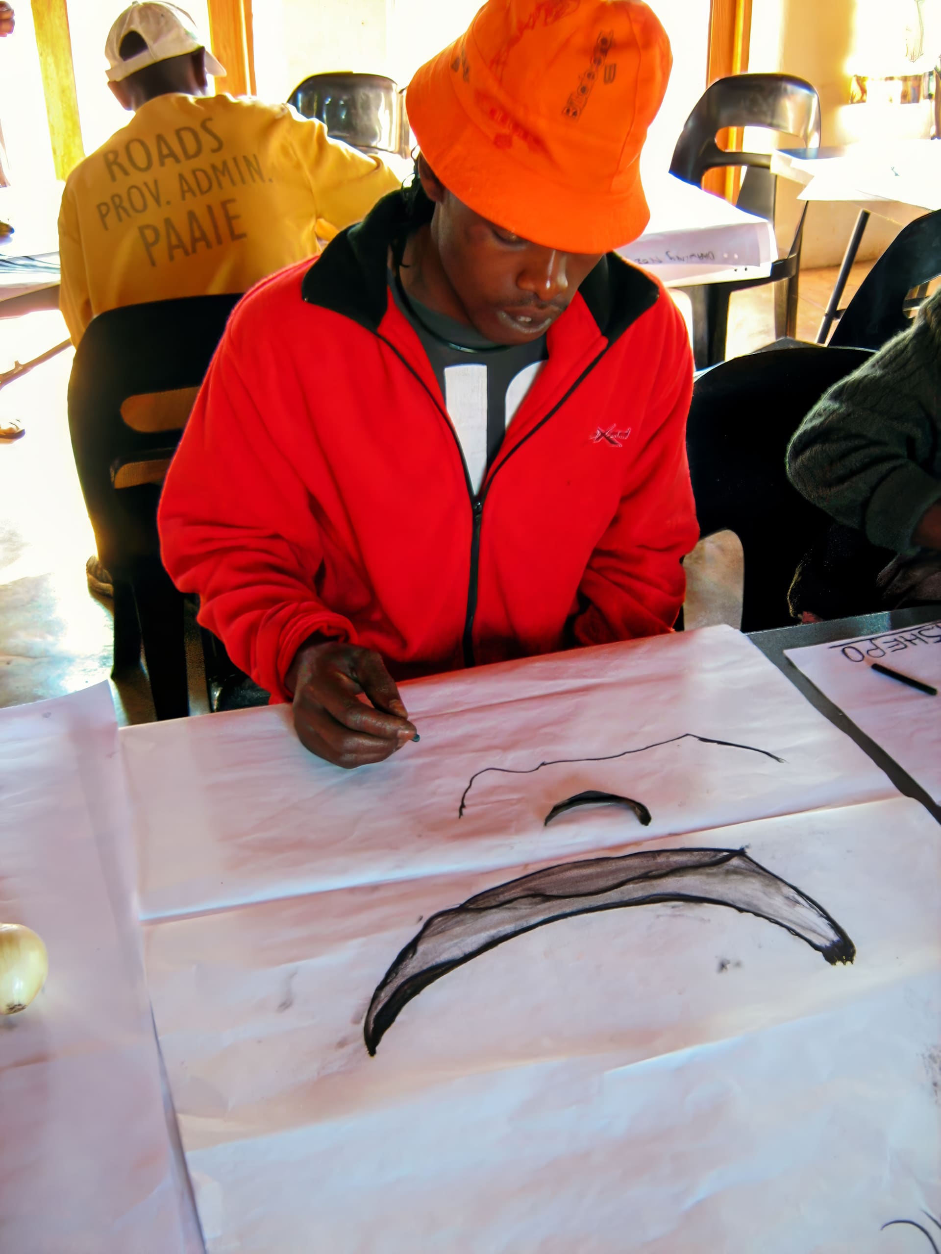 A person in an orange hat and red jacket sits at a table, drawing two dark, curved shapes on a large sheet of white paper. In the background, another person in yellow faces away, with “ROADS PROV. ADMIN PAARLE” visible on their shirt.