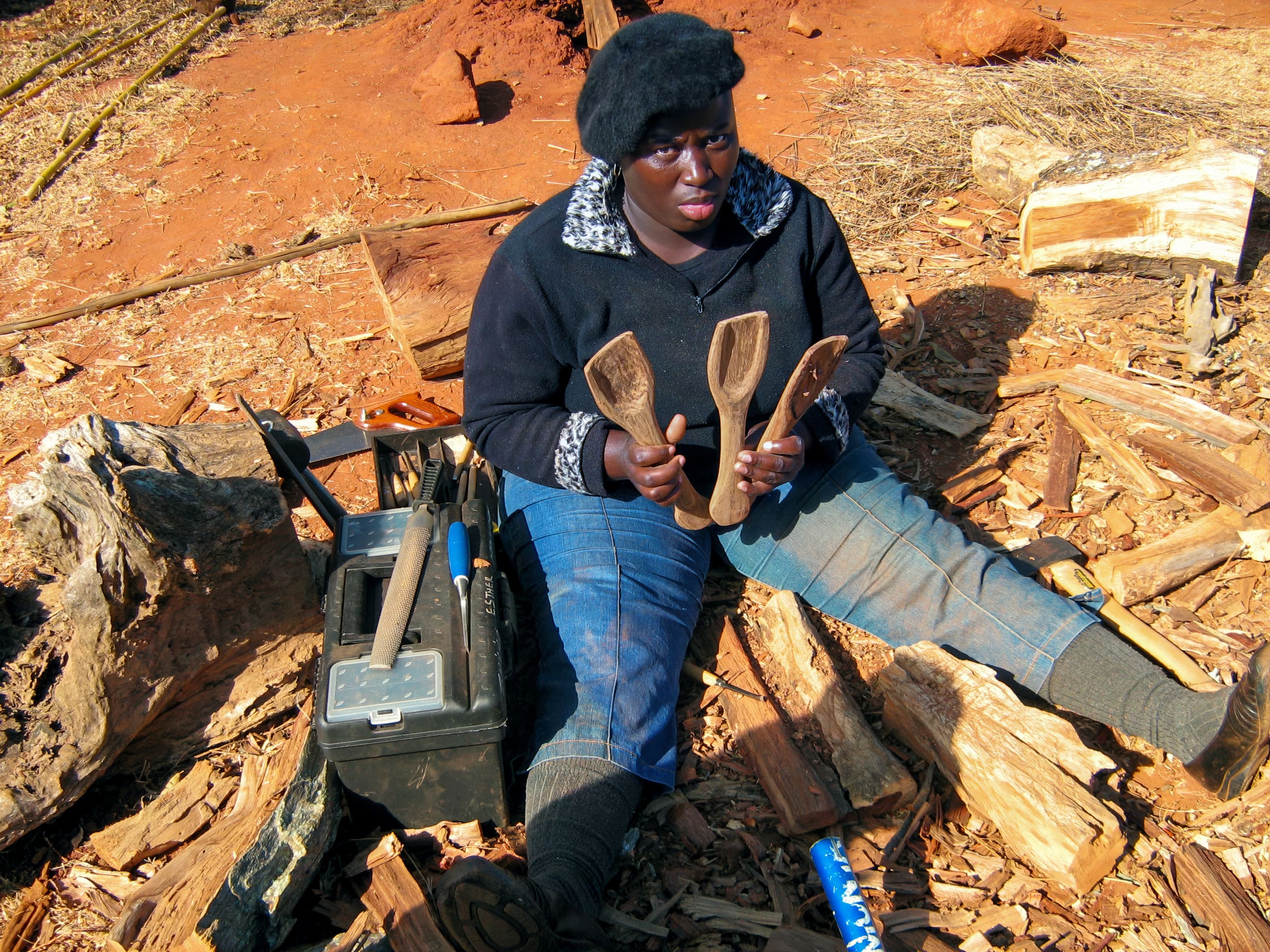 A woman in a black hat and blue jeans sits on the ground among wood shavings, holding three hand-carved wooden spoons. A toolbox with carving tools is beside her, and chopped logs and pieces of bark are scattered around on reddish soil.