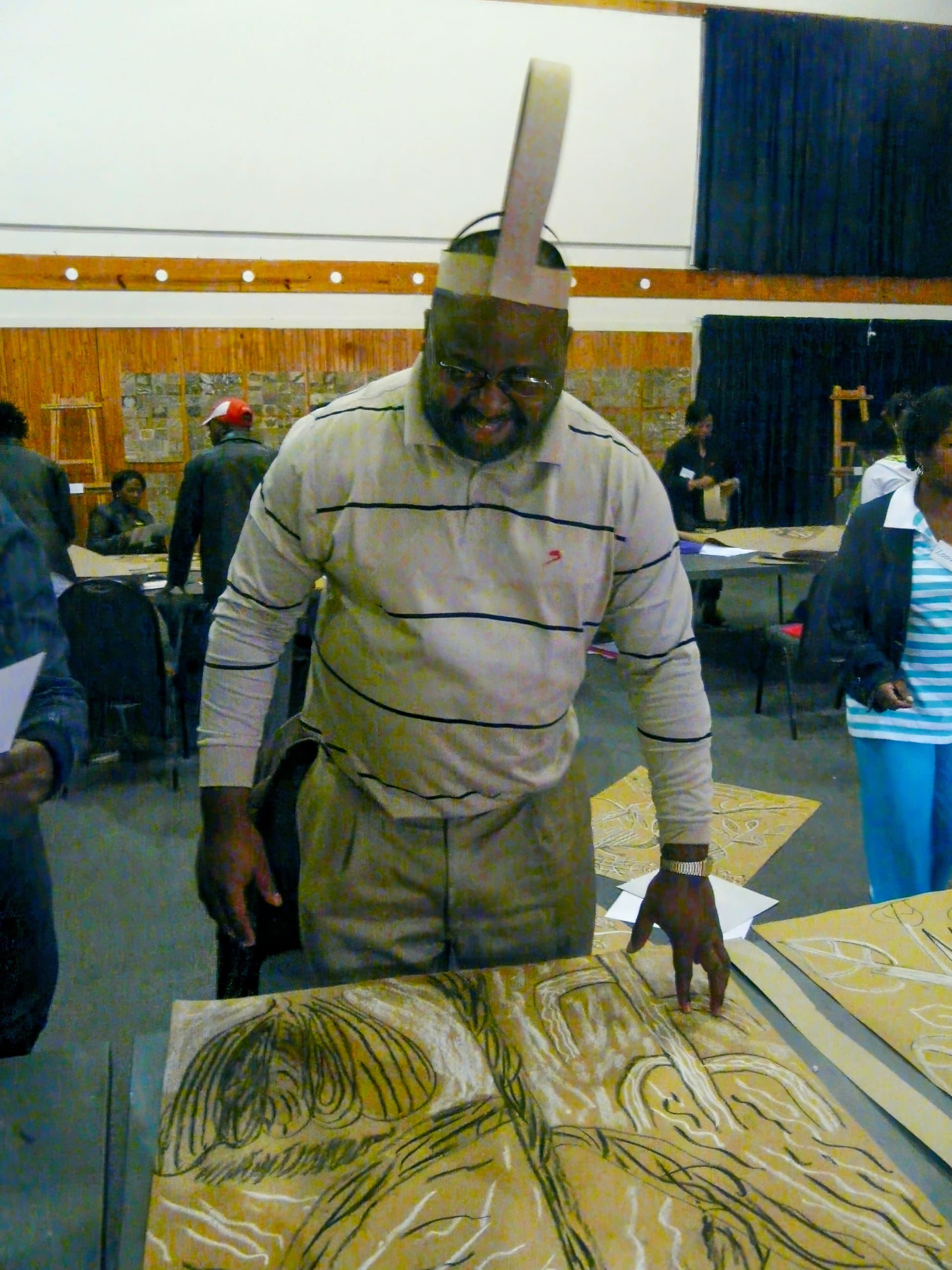A man wearing glasses and a striped jumper stands indoors, smiling and looking down at a carved wooden panel. He has a strip of paper bent into a loop and attached to his head. Other people are gathered around tables in the background, engaged in similar activities.