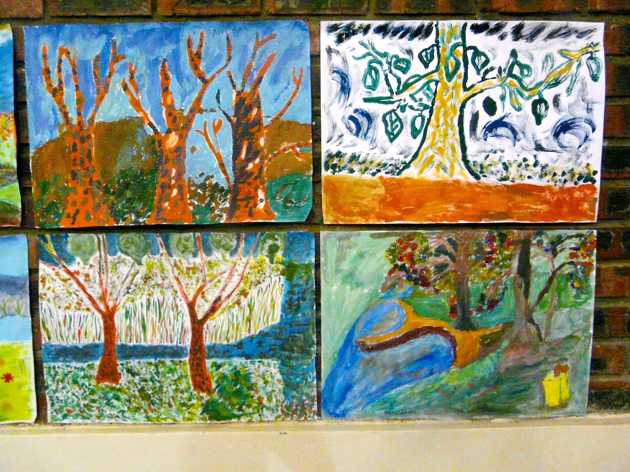 Four colourful paintings of trees are displayed on a brick wall. Each painting shows stylised trees with different backgrounds and colours, including blue skies, orange trunks, green leaves, and a river. The styles vary, appearing playful and expressive.