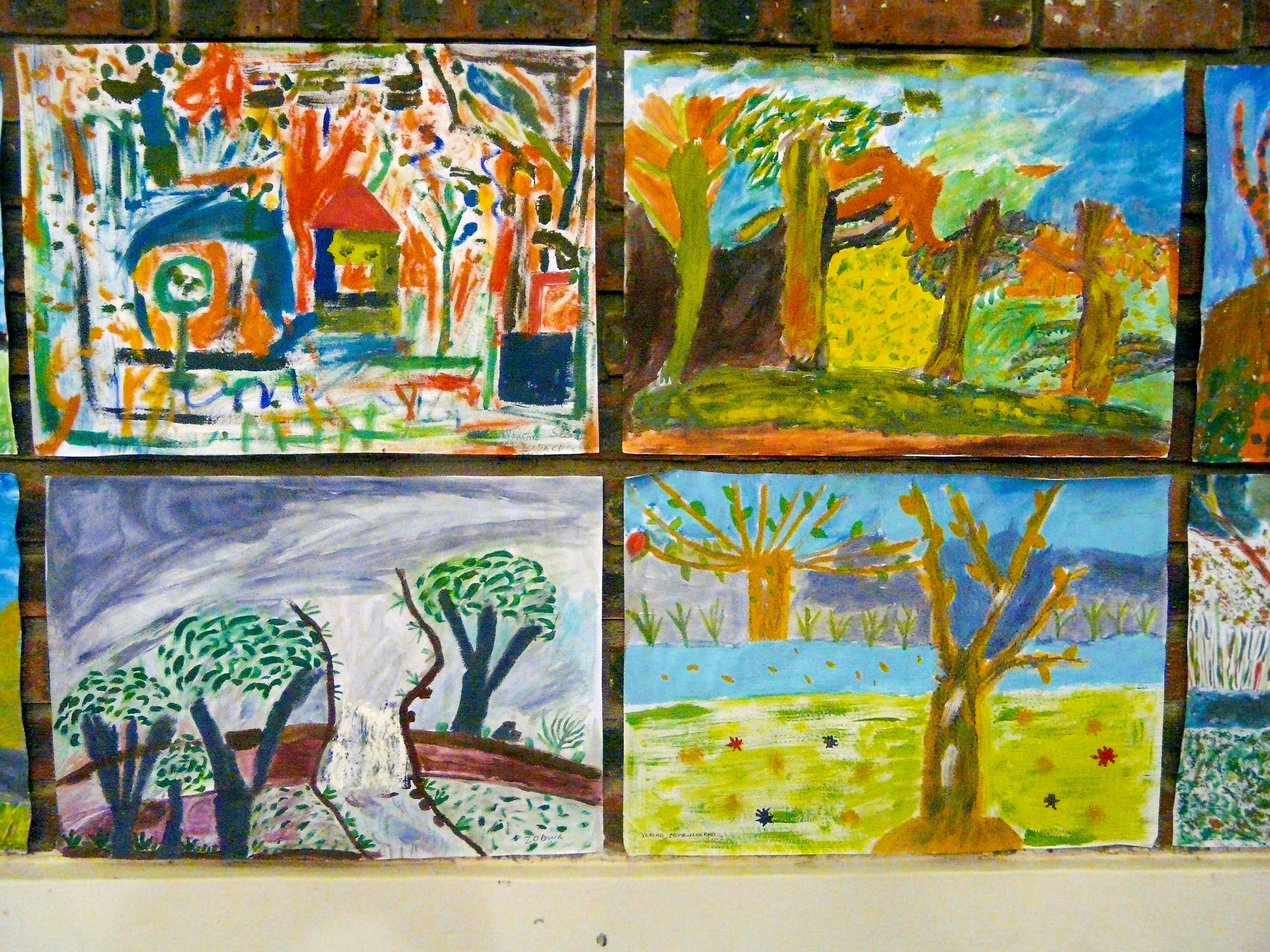 Four colourful, expressive paintings are displayed on a wall. Each depicts a vibrant outdoor scene with trees, grass, and varying skies, using bold brushstrokes and bright colours. The artworks appear to be created by children or beginners.