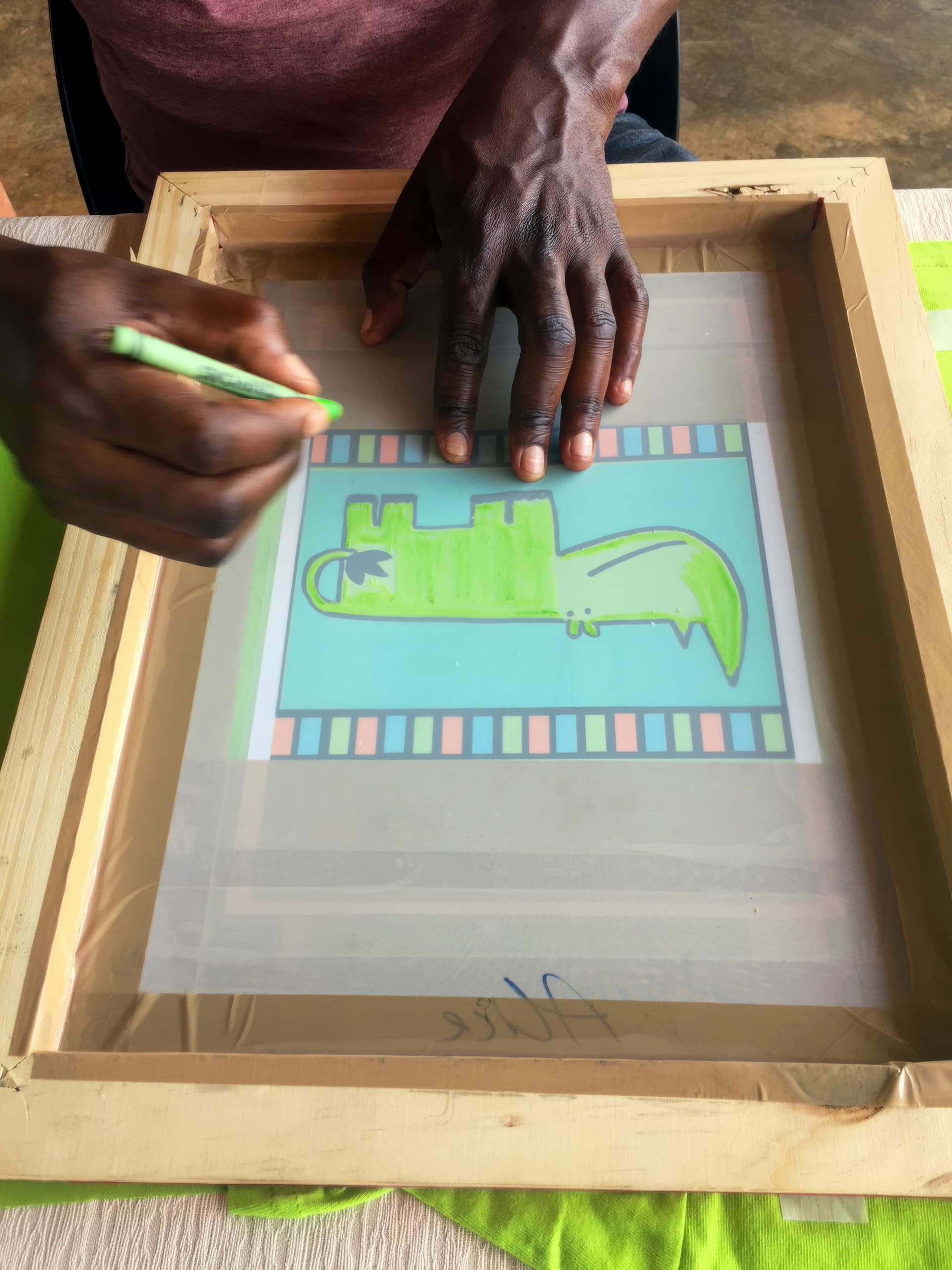 A person’s hands trace a green cartoon crocodile onto a clear screen for printmaking. The drawing is framed by a wooden screen and features a blue background with a colourful, striped border. The person uses a green pencil, with fabric visible underneath the frame.