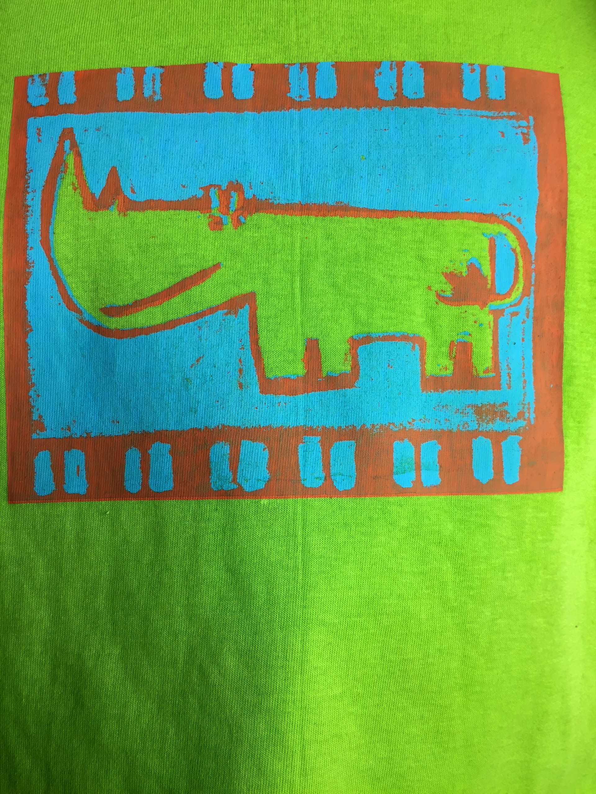 A stylised orange rhino with a long horn is outlined within a blue rectangular frame, surrounded by small blue rectangles, all set against a bright green fabric background. The design has a playful, hand-drawn cartoon appearance.