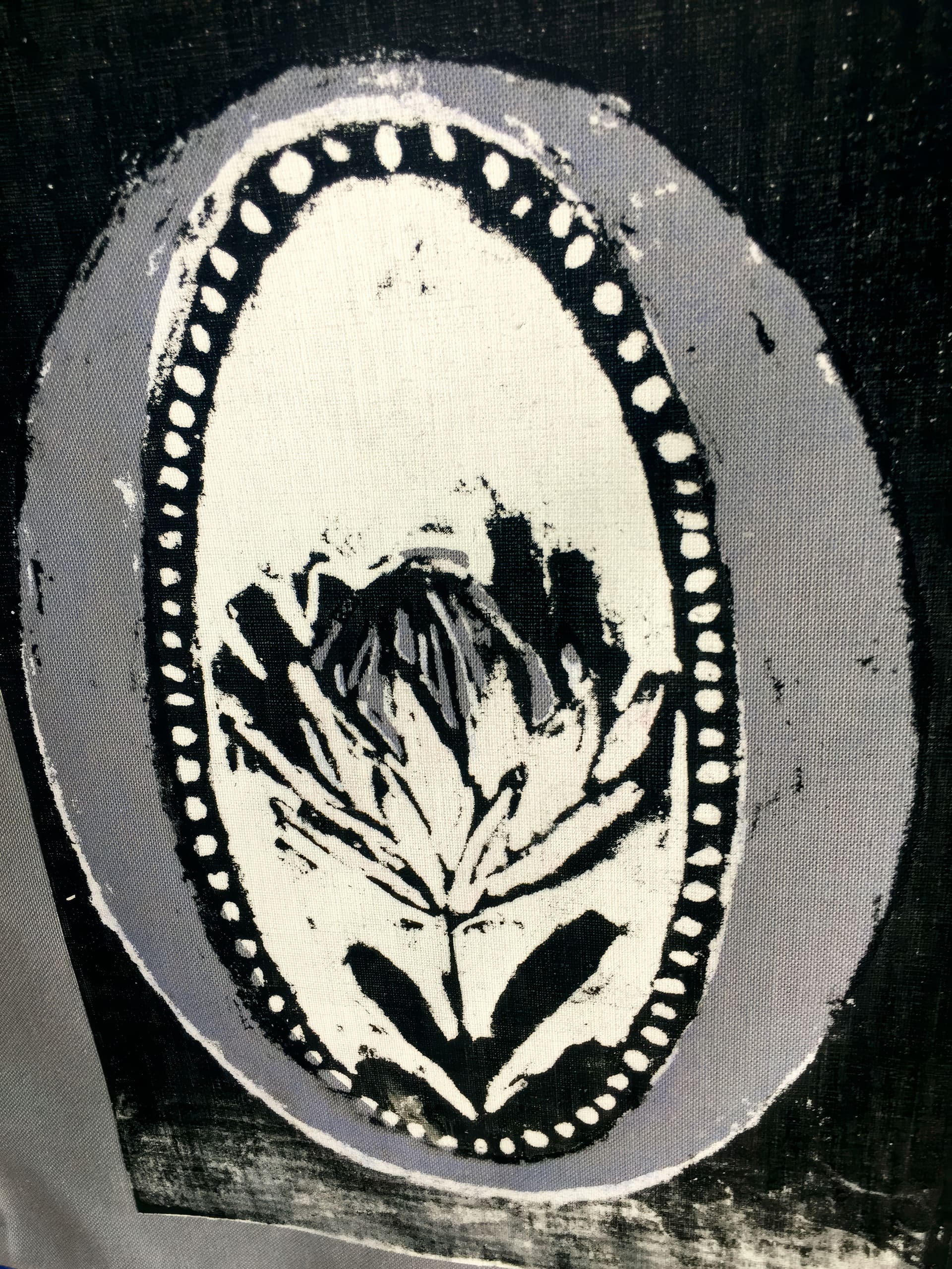 A black and white fabric print features an oval frame with a dotted border. Inside the oval, there is a stylised, abstract flower with bold, dark brushstrokes on a light background. The outer edges are shaded in grey, giving a textured appearance.