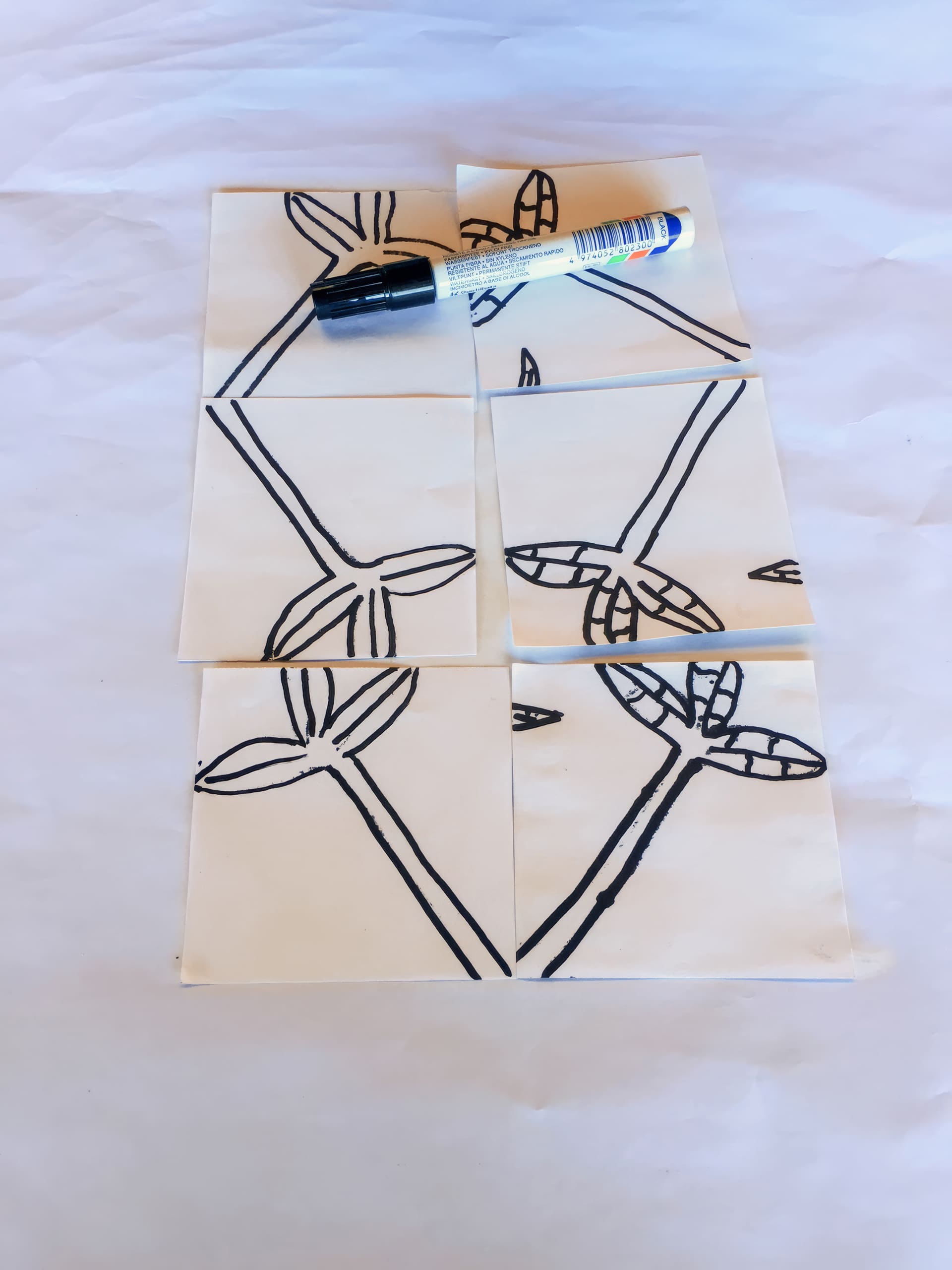 Six square papers with parts of a black marker line drawing of leaves and stems are arranged together to form a complete image. A black-capped marker is placed on top of the upper centre square. The background is a white, slightly wrinkled surface.