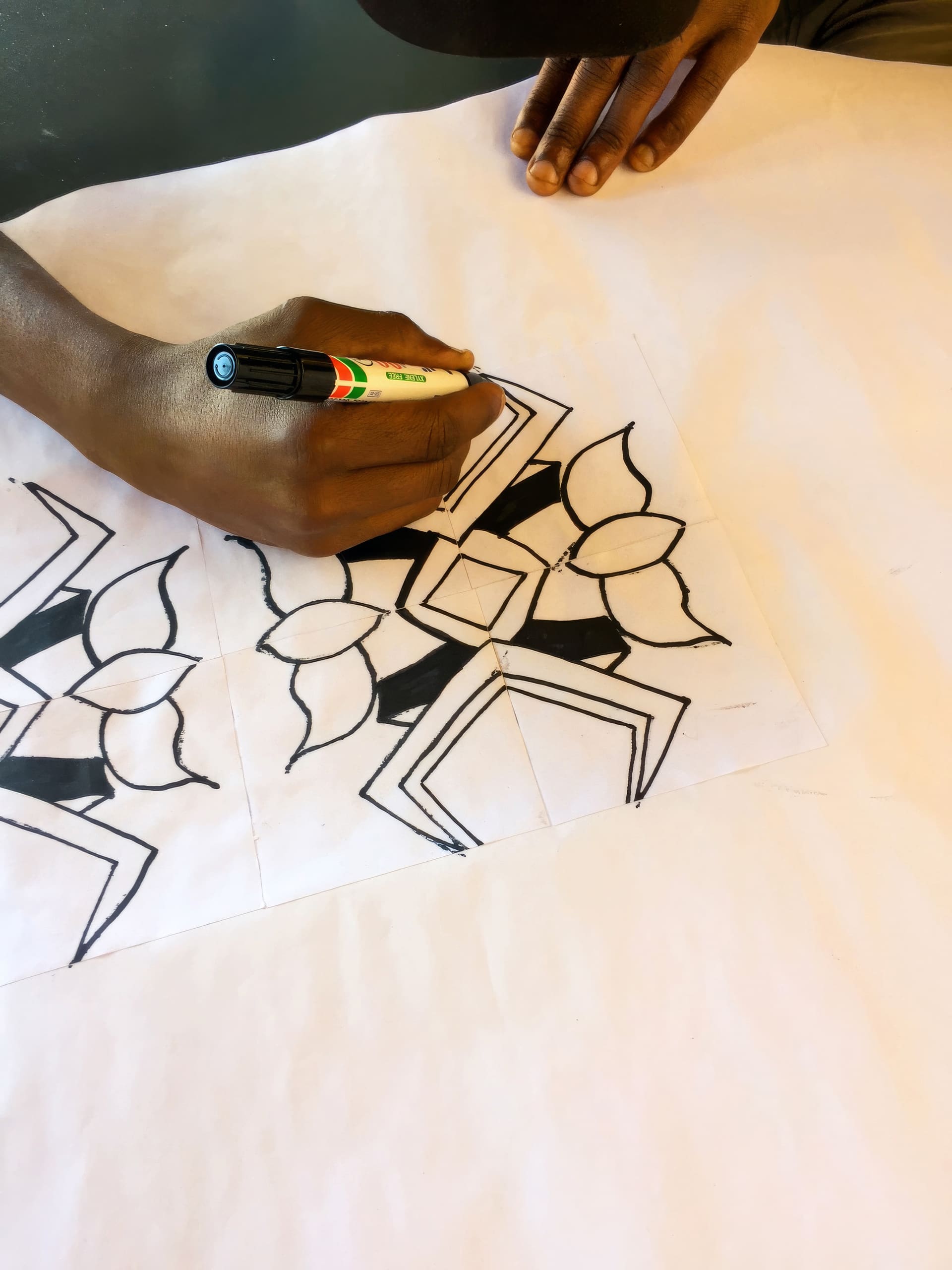 A person’s hand draws geometric and floral patterns with a black marker on a white sheet. The design features repeated hexagons and petal shapes. Another hand rests nearby on the paper, and the background is out of focus.
