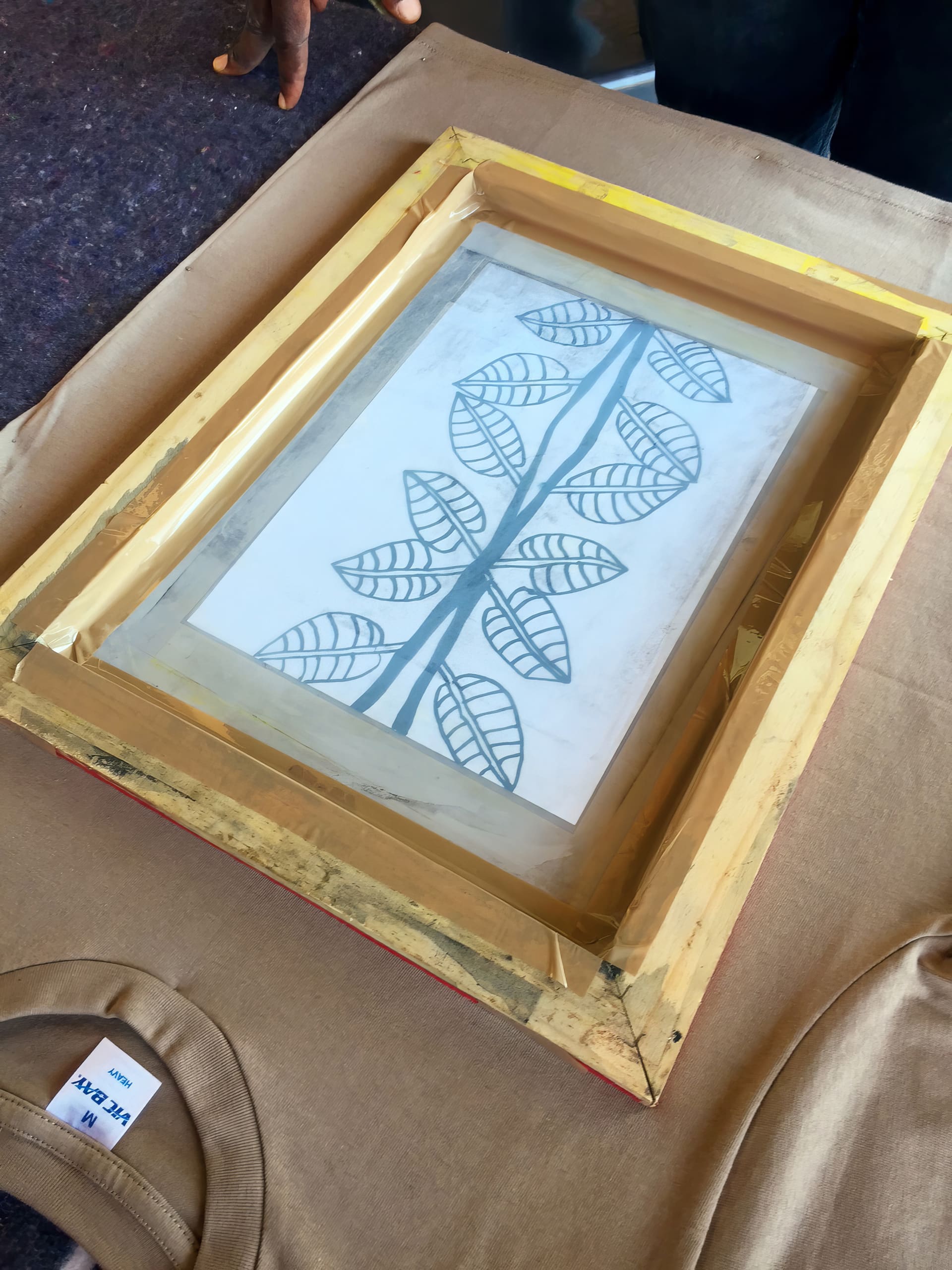 A wooden screen-printing frame with a transparent sheet showing a leaf pattern design lies on a beige surface. Part of a tan T-shirt with a visible size label is in the foreground, and a person's hand appears at the top left corner.