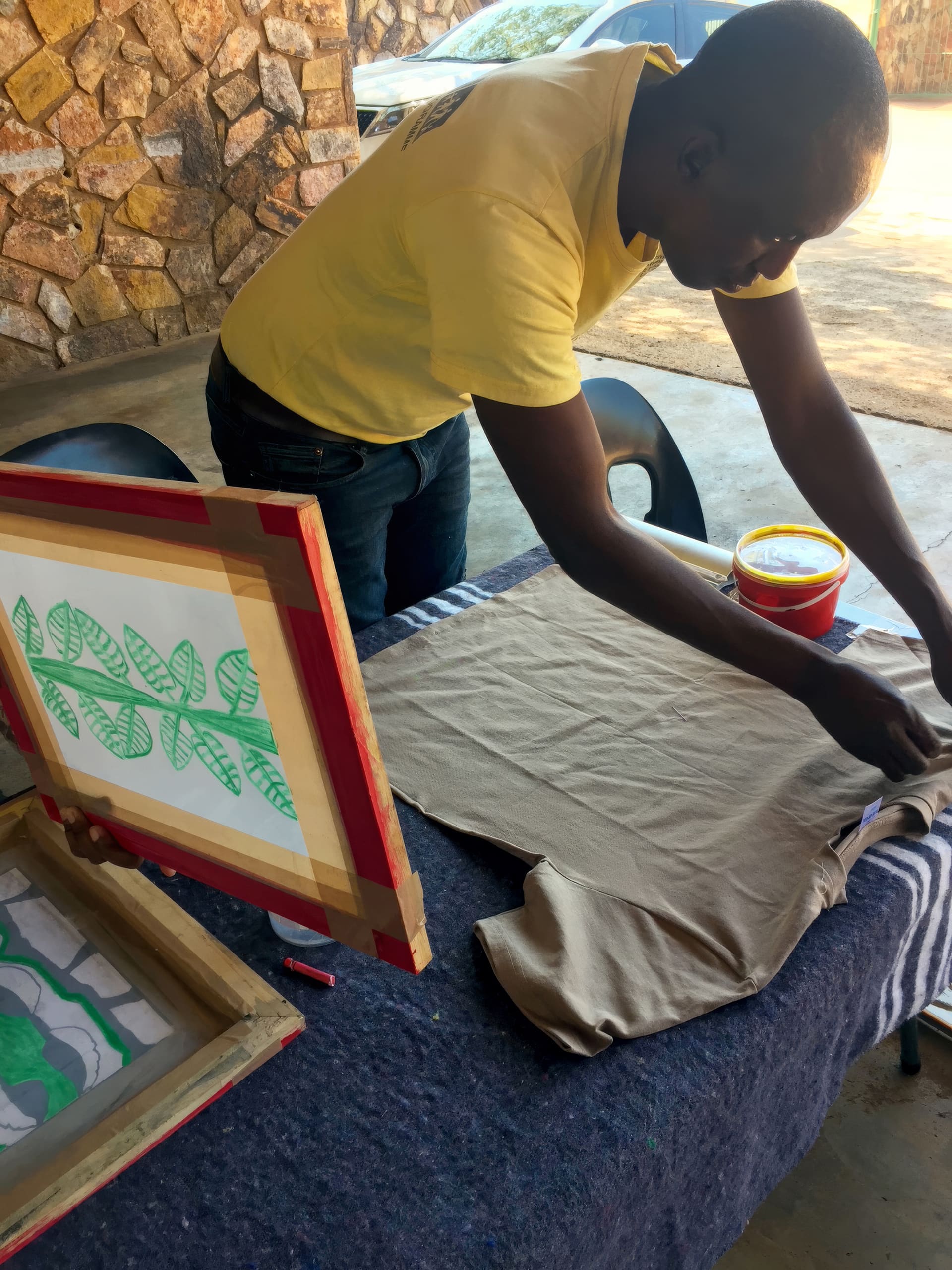 A person in a yellow shirt prepares a beige T-shirt for screen printing on a table outdoors. Two screens with leaf designs, a container of yellow paint, and a stone wall are visible in the background. The person is focused on arranging the shirt.