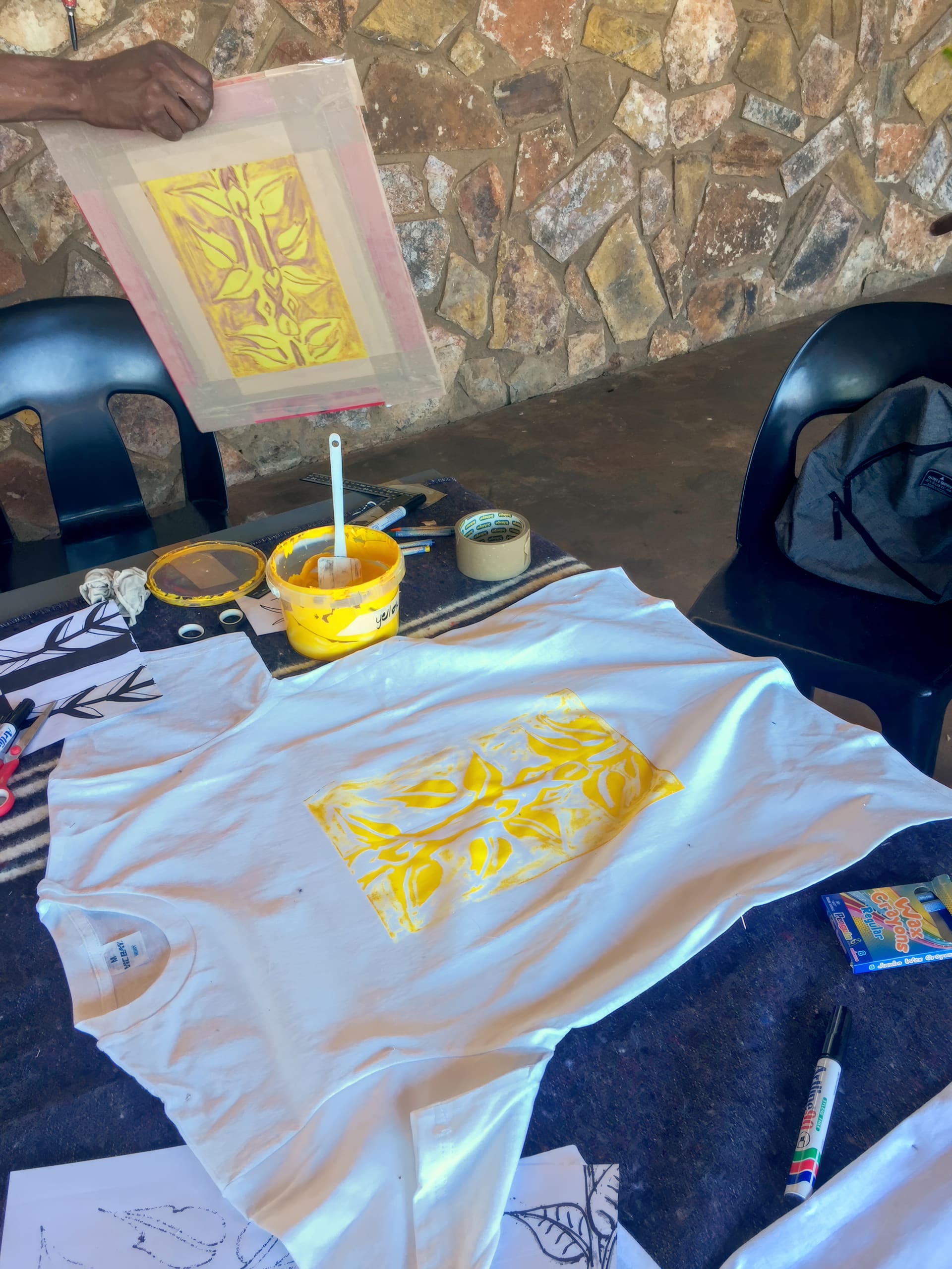A hand holds a screenprinting frame above a table with a white T-shirt that has a yellow patterned print in the centre. Nearby are a paintbrush in a yellow paint container, tape, markers, and drawing supplies on a dark cloth. A stone wall is in the background.