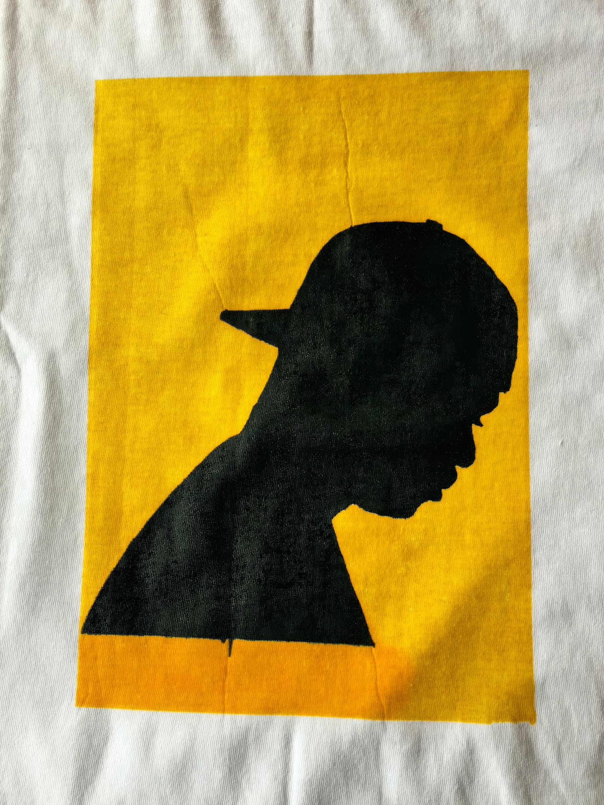 A black silhouette of a person in profile view wearing a baseball cap, facing right, is shown against a solid yellow rectangular background on a white surface. The image has a bold, graphic style.