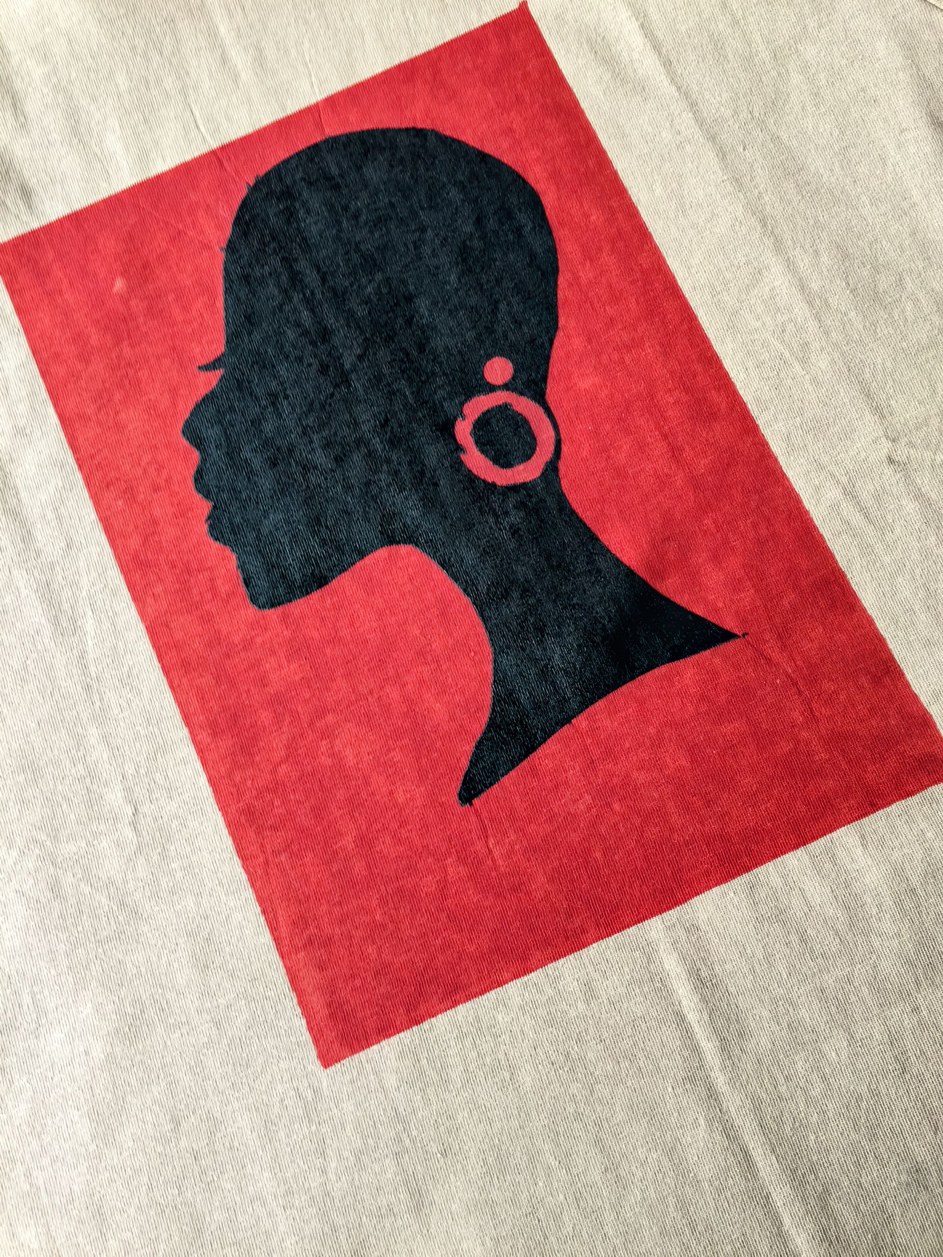 A black silhouette of a woman’s head in profile, with a large hoop earring, is centred in a solid red rectangle on a beige fabric background. The woman’s features are simple and stylised, with visible eyelashes and a short hairstyle.