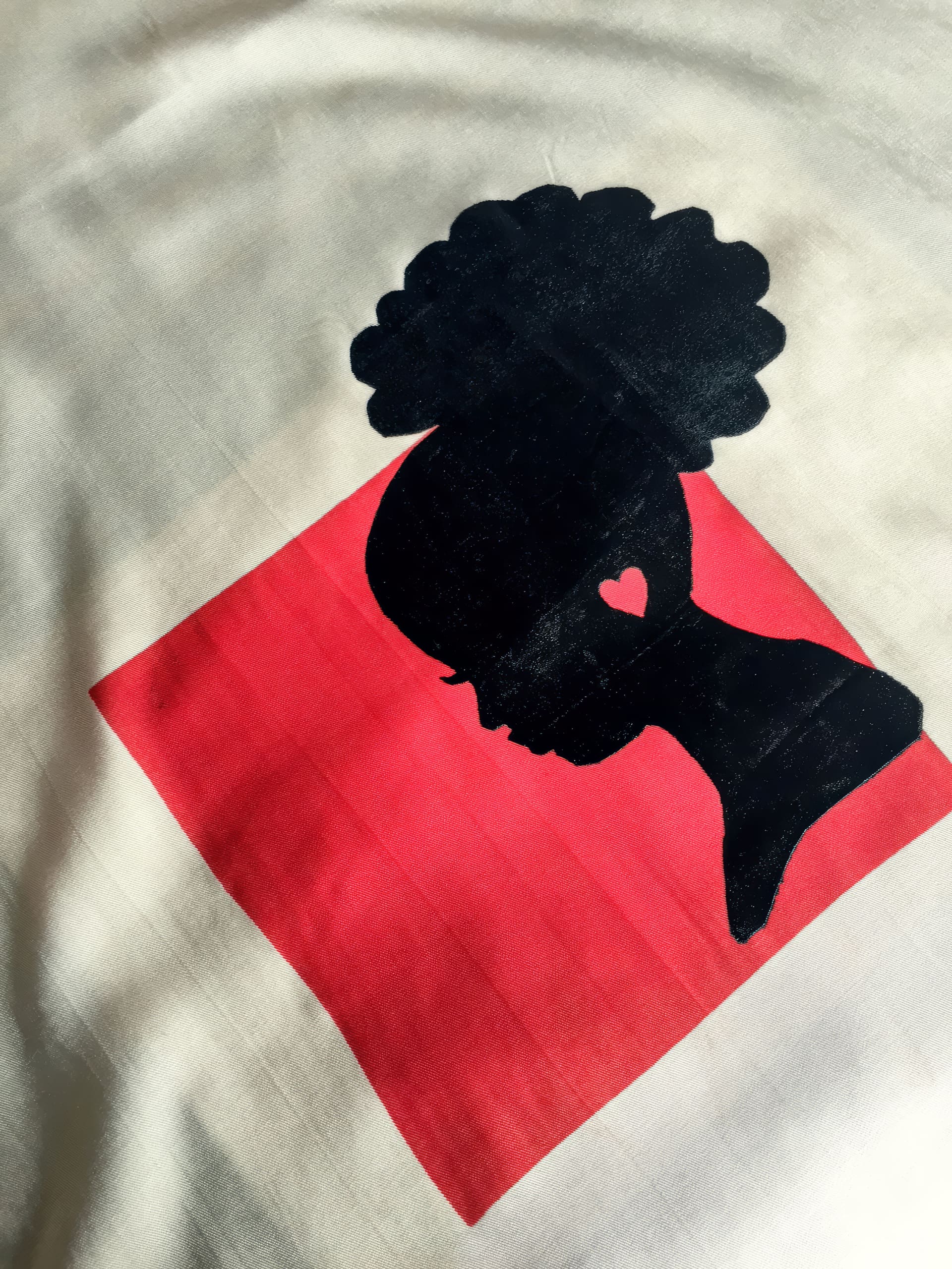 A black silhouette of a woman with an afro and a heart shape on her cheek is centred over a red square background on a cream-coloured fabric. The woman is shown in profile, facing left, with elegant facial features.