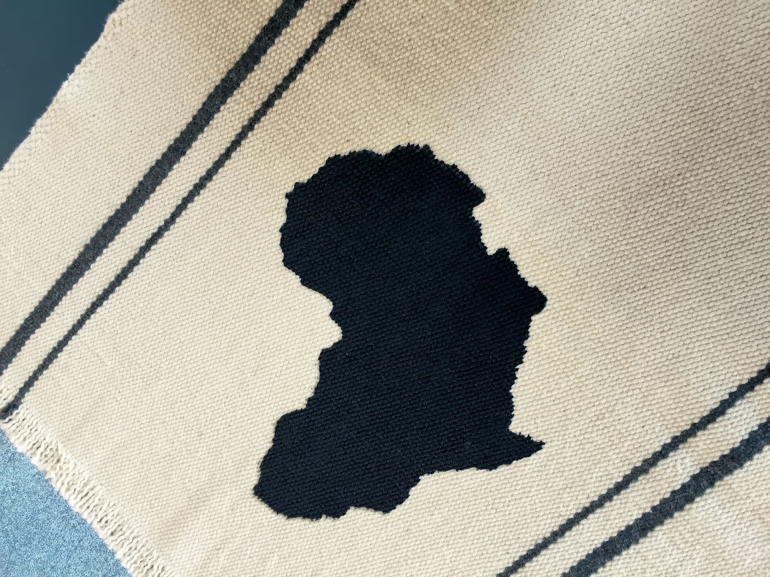 A beige woven rug features a large, irregular black shape in the centre resembling a silhouette or inkblot, with two parallel black stripes running diagonally along the sides of the rug. The texture of the weave is visible.